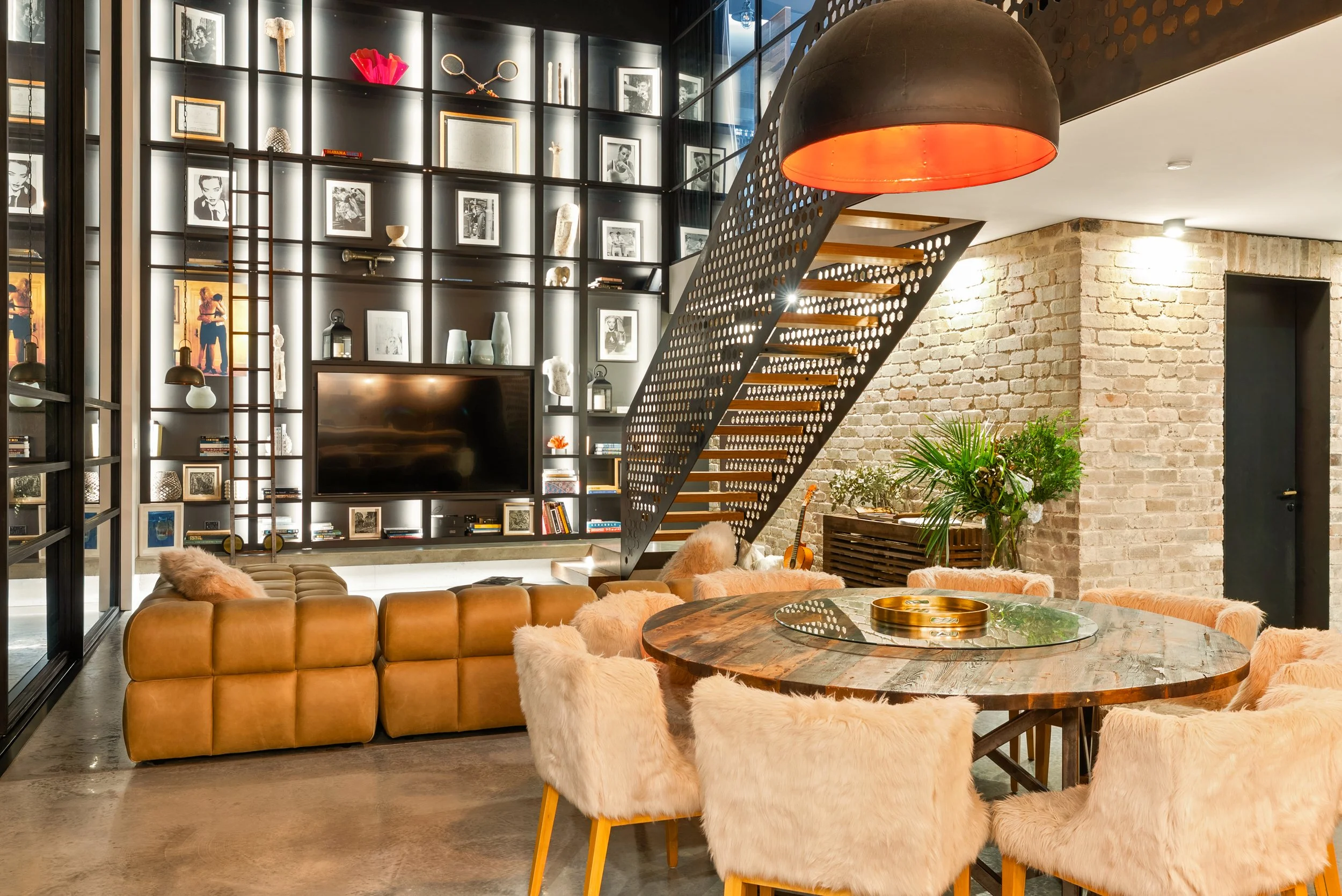 Modern living room with a round wooden dining table surrounded by fluffy beige chairs, a sectional couch, a black wall of shelves with pictures and decorative items, a staircase with perforated metal sides, and a brick wall painted in warm tones.