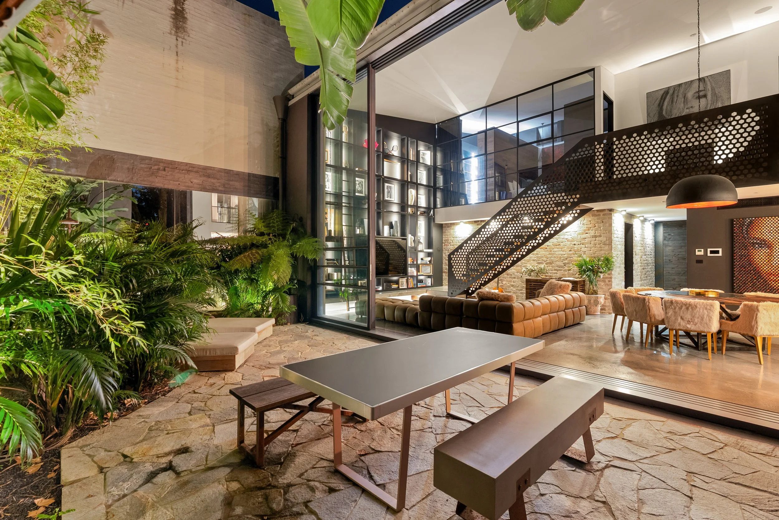 Modern living room with a staircase, large window, and a mix of indoor and outdoor seating areas, decorated with plants and contemporary furniture.