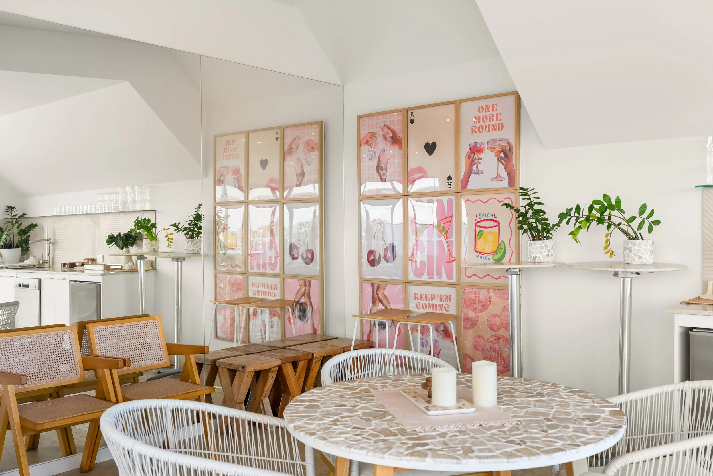 Decorative pink and red-themed posters on a glass partition in a bright, minimalist room with white walls, potted plants, wooden chairs, and a round table with candles.