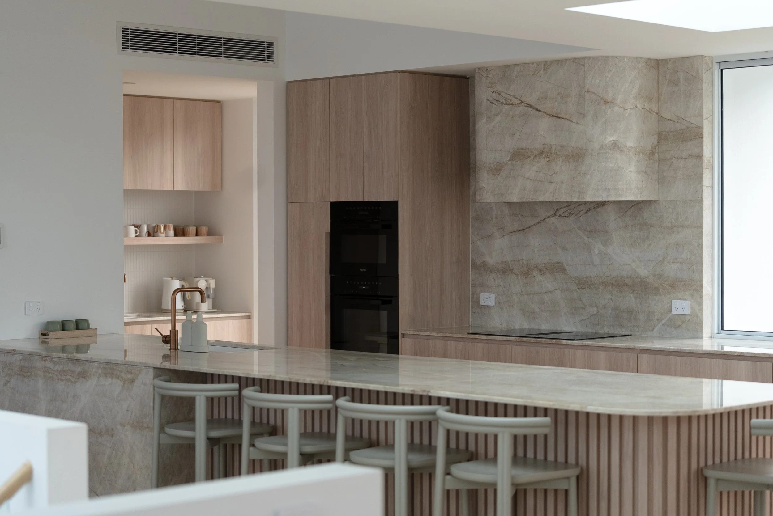 Modern kitchen with beige marble countertops, light wood cabinetry, and a wall of large marble tiles. Bar stools are lined up along the island, and there is a window on the right side.