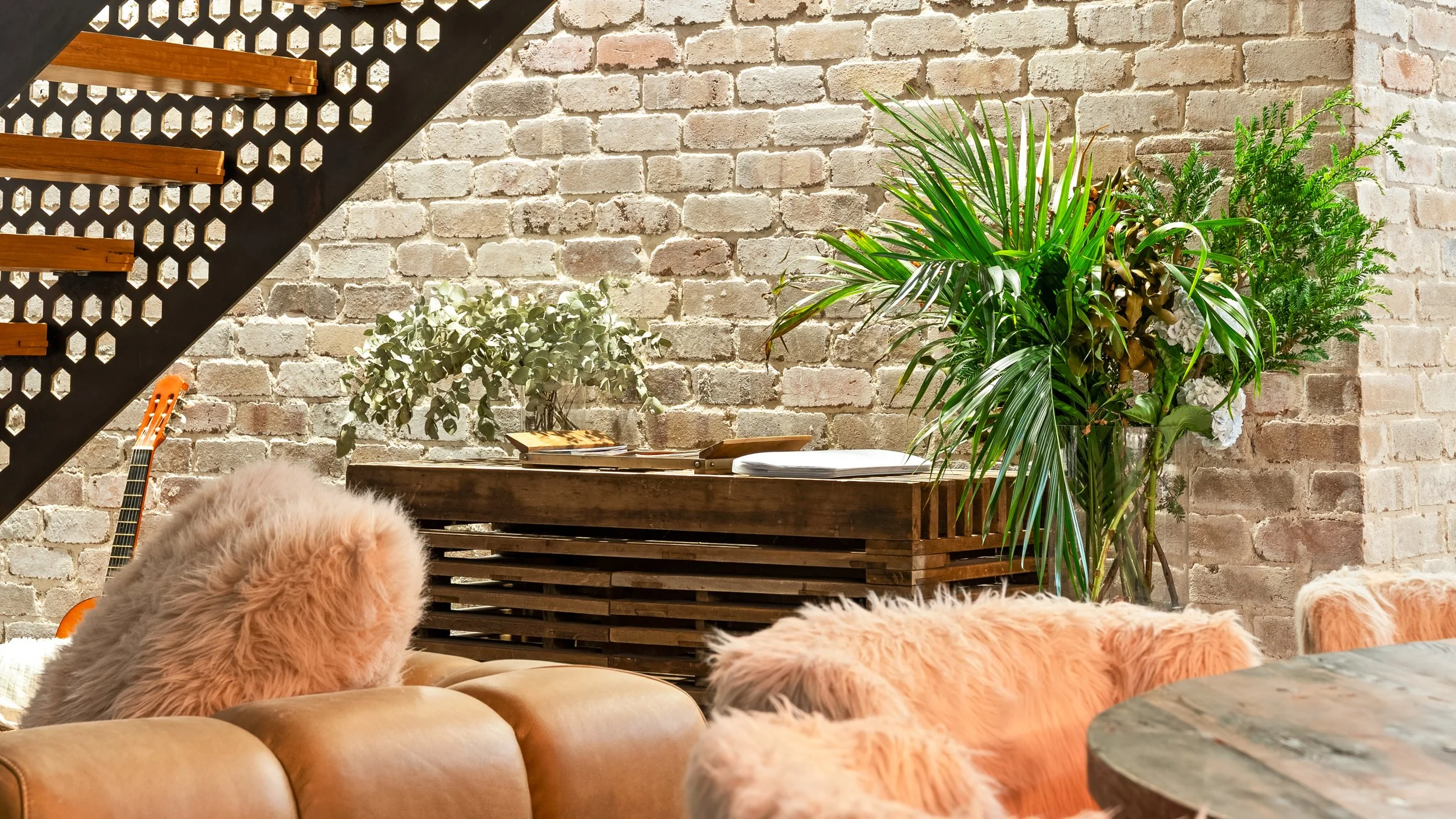 Living room with a brick wall, large green plants, a wooden sideboard, and a cozy couch with pink furry pillows.