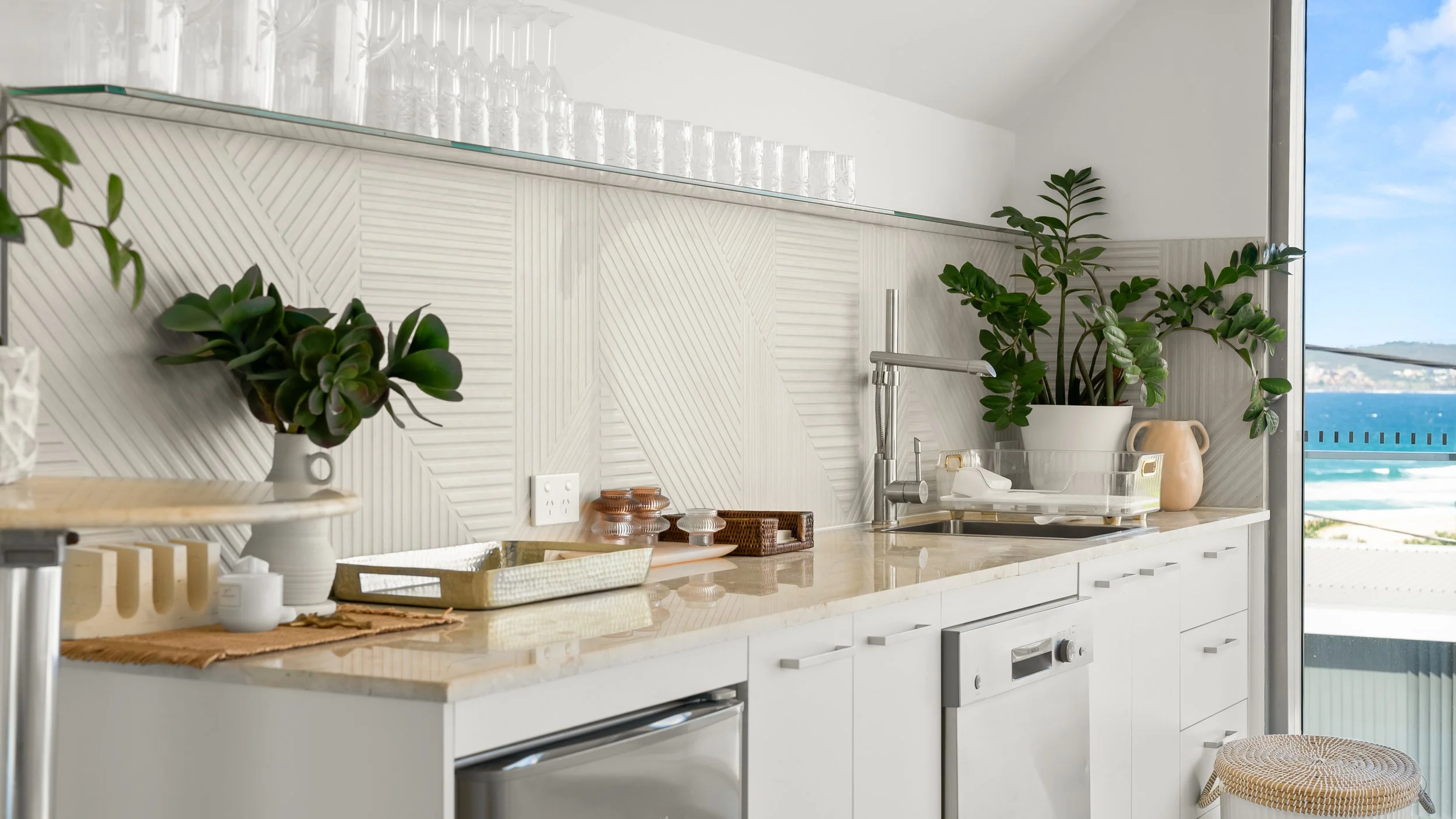 Modern kitchen with white cabinetry, a marble countertop, and potted plants near a large window overlooking a view of the ocean and blue sky.