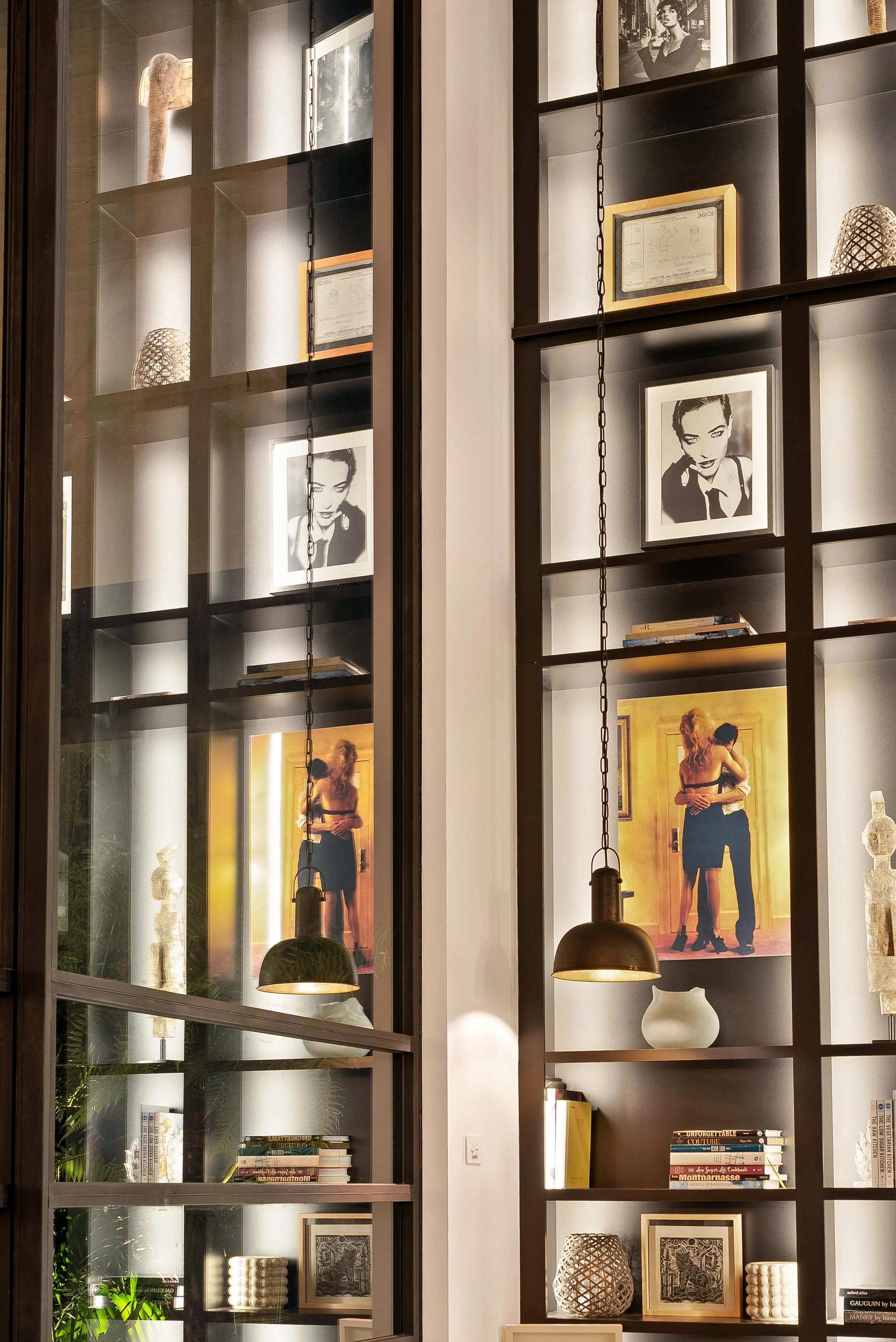 Luxury interior with built-in black shelves displaying framed photographs, artwork, decorative objects, and books, illuminated by built-in lighting and pendant lights.