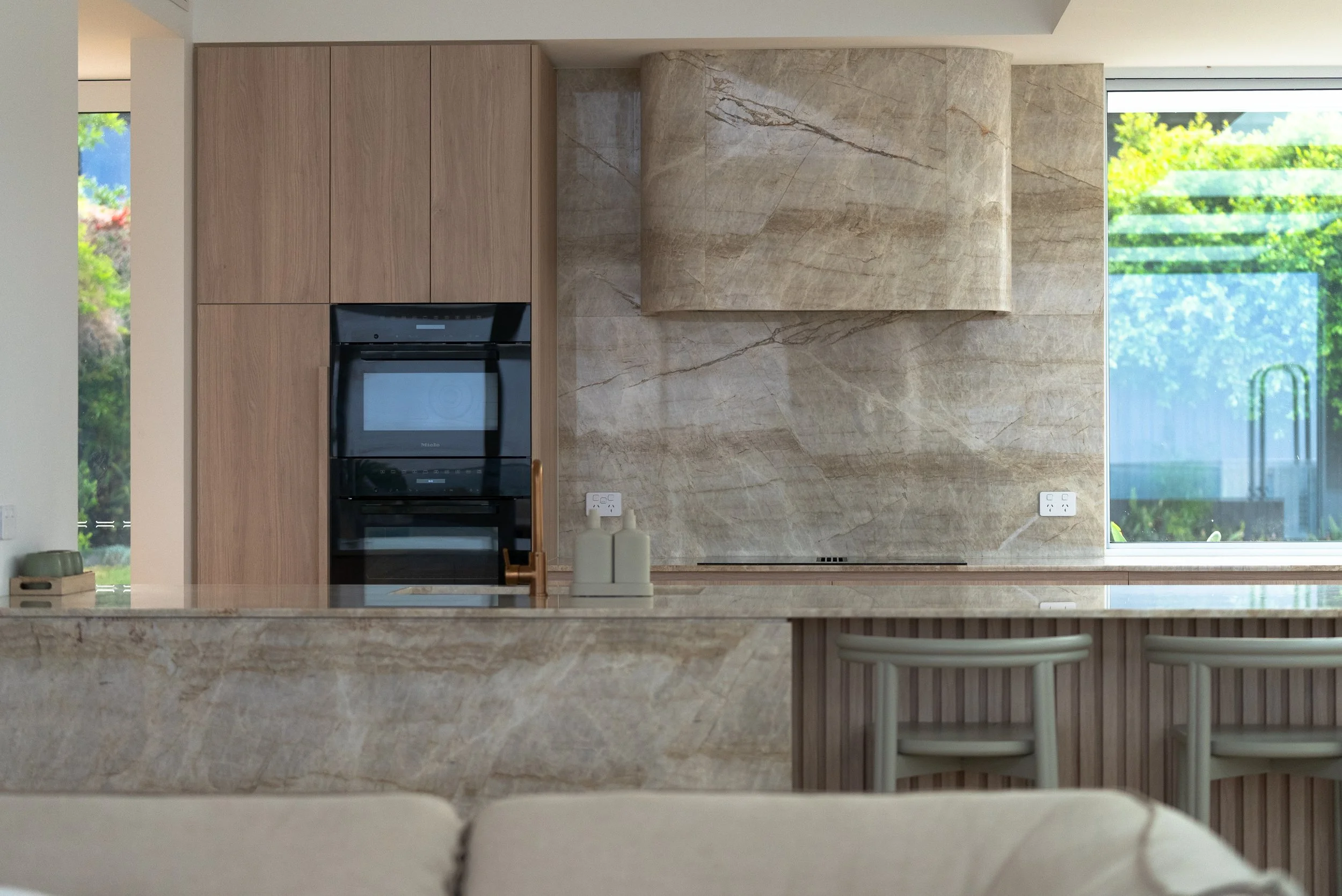 Modern kitchen with beige marble backsplash, wood cabinets, and a built-in black oven with a microwave, adjacent to a large window showing greenery outside.