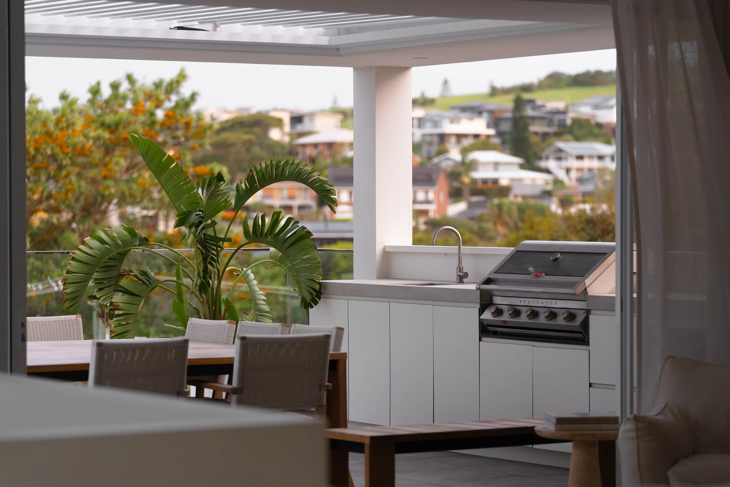 Outdoor balcony with a dining table, chairs, a large potted plant, a grill, sink, and a scenic view of houses and greenery in the background.