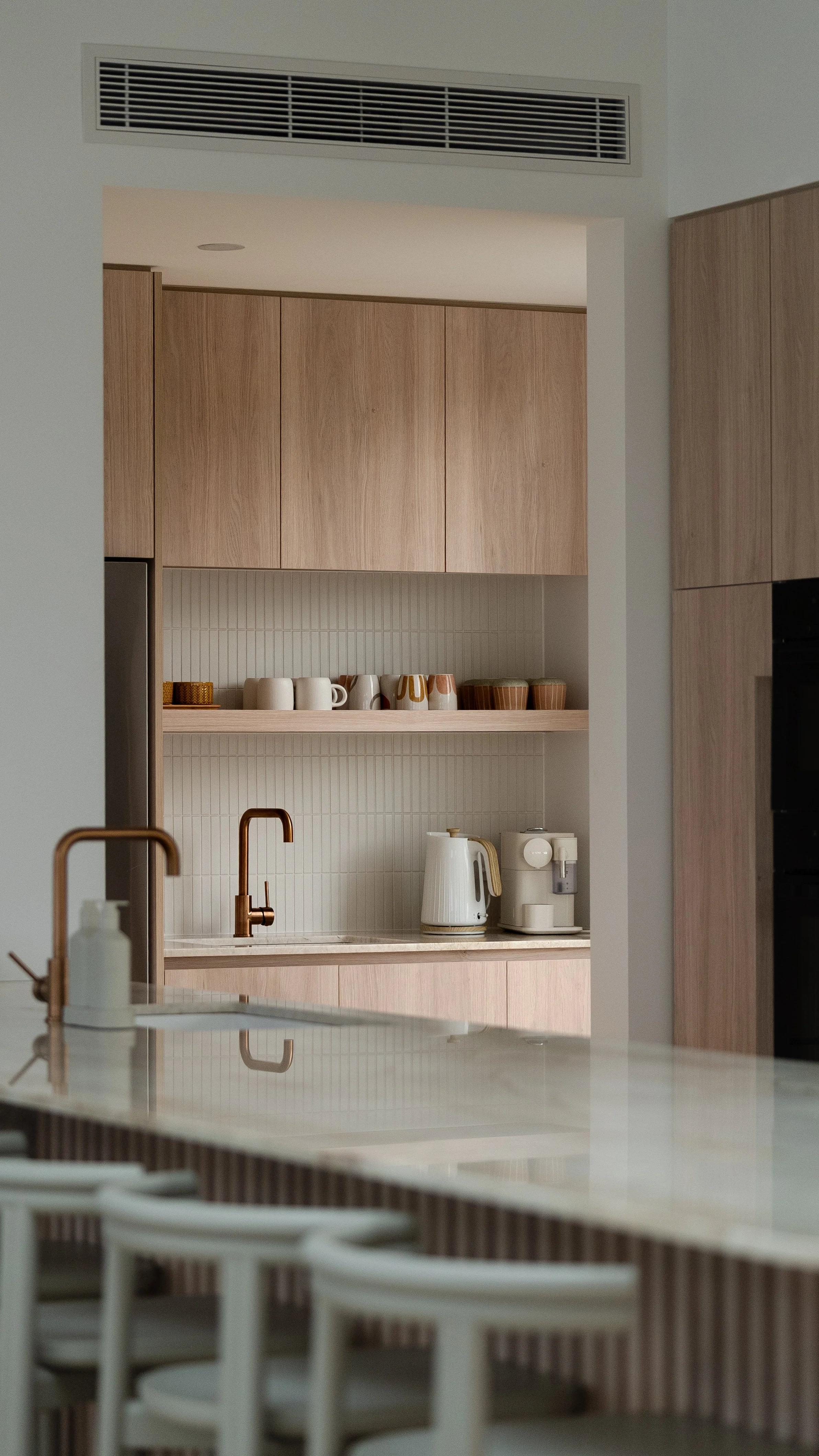 Modern kitchen with light wood cabinets, a white countertop, a copper faucet, and a shelf with cups and bowls. There is a white electric kettle and a white coffee machine on the counter.