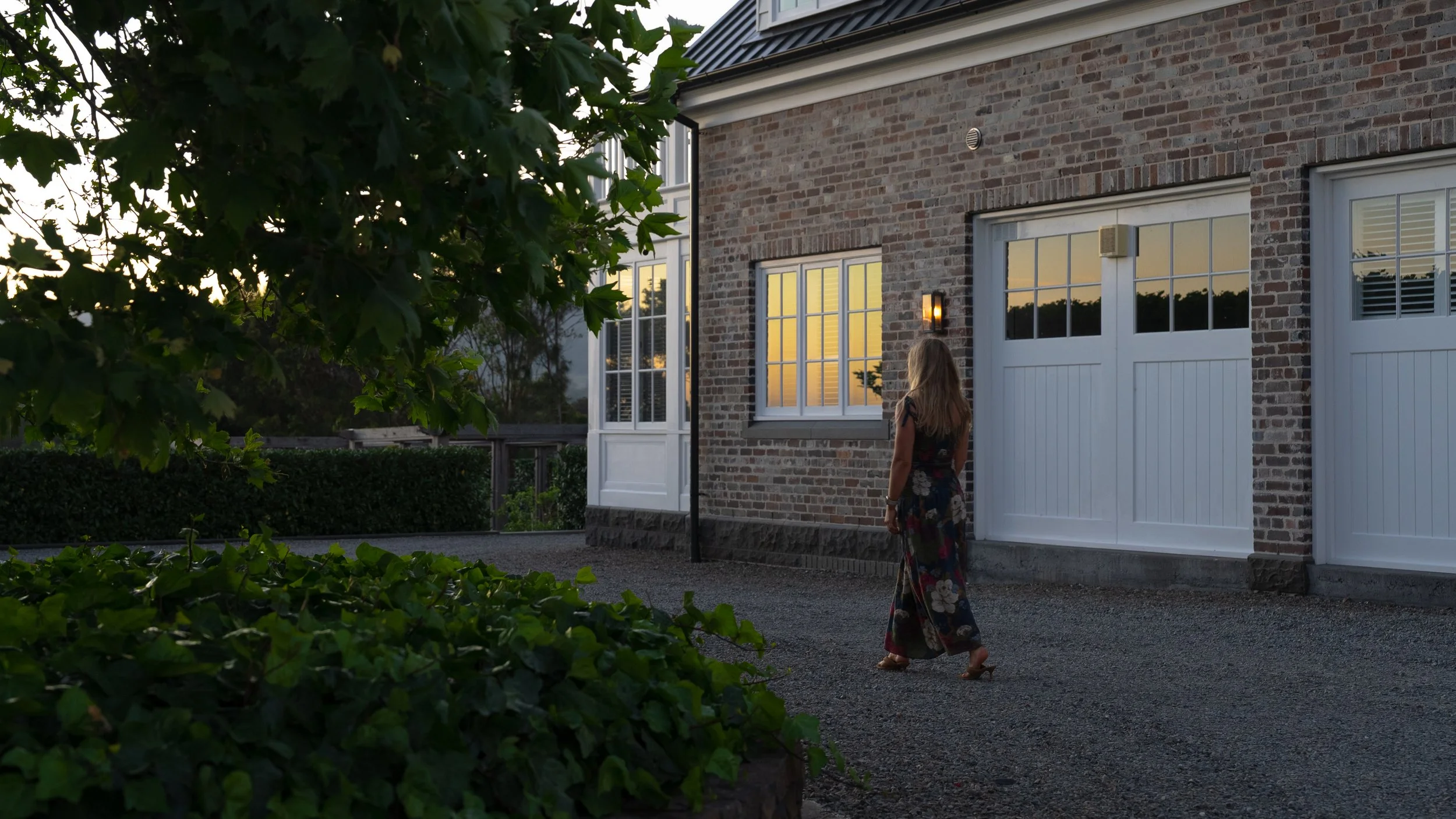 A woman in a floral dress walking towards a brick house with white garage doors and windows, during sunset, with greenery in the foreground.
