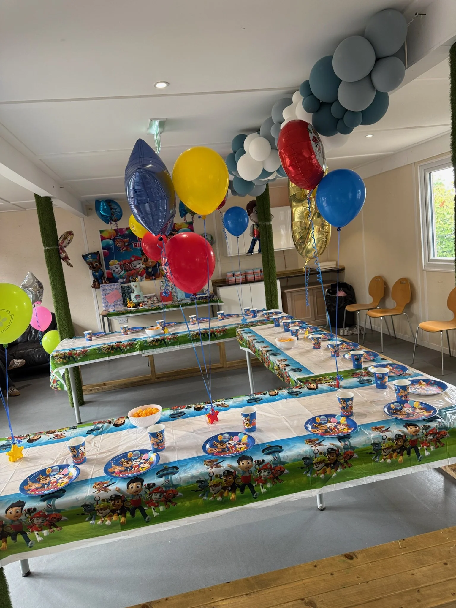 A decorated indoor party setup featuring tables covered with colorful Paw Patrol themed tablecloths, surrounded by balloons in various colors including red, blue, yellow, and silver. There is a blue and gray balloon arch on the ceiling, and paper cups and plates with Paw Patrol characters are arranged on the tables. The room has beige walls, a window on the right, and some chairs against the wall.