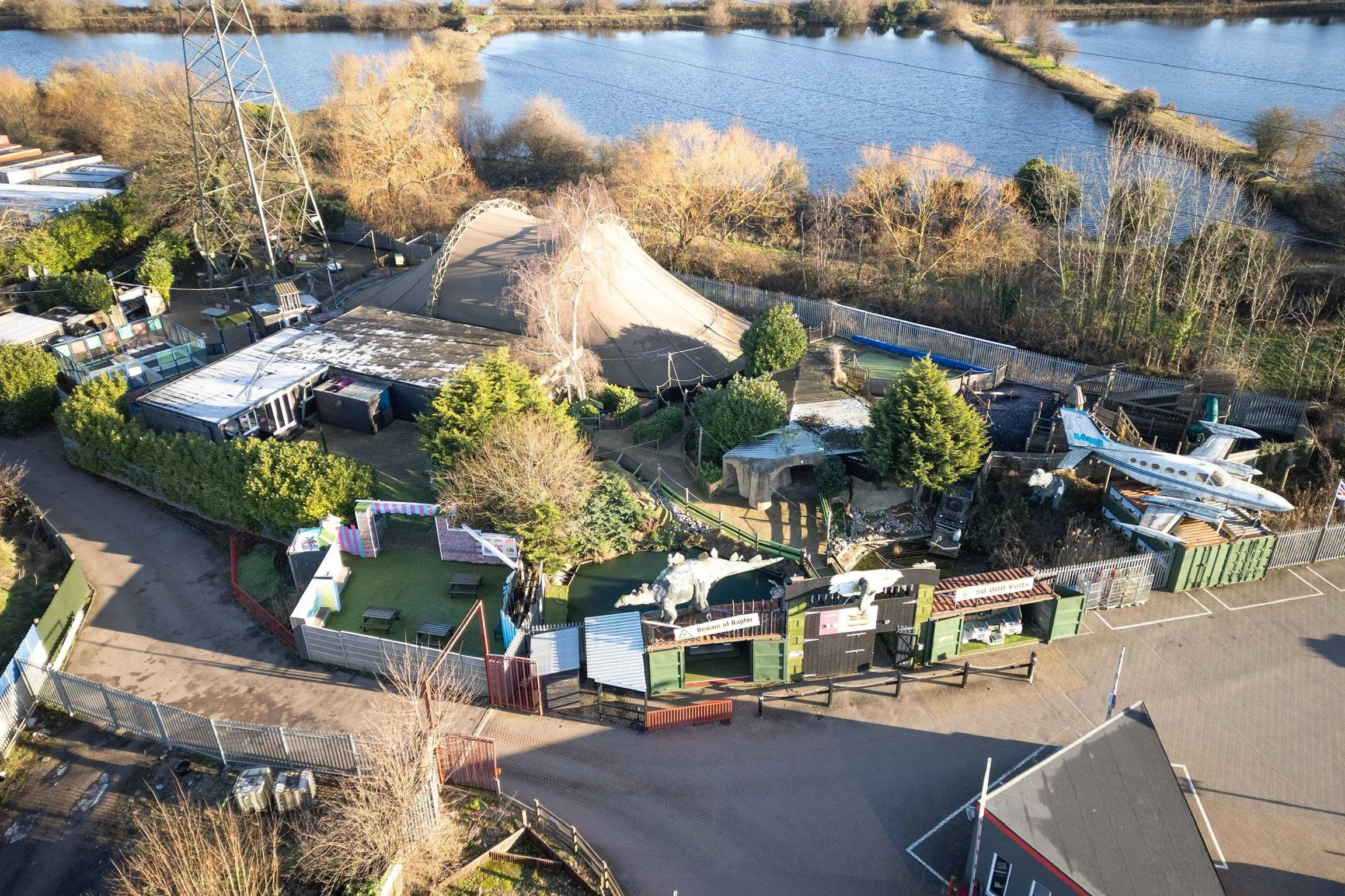 Aerial view of a small amusement park enclosed by a fence, featuring a miniature airplane ride, a tent, a pond, and landscaped areas, with trees, water, and a parking lot nearby.