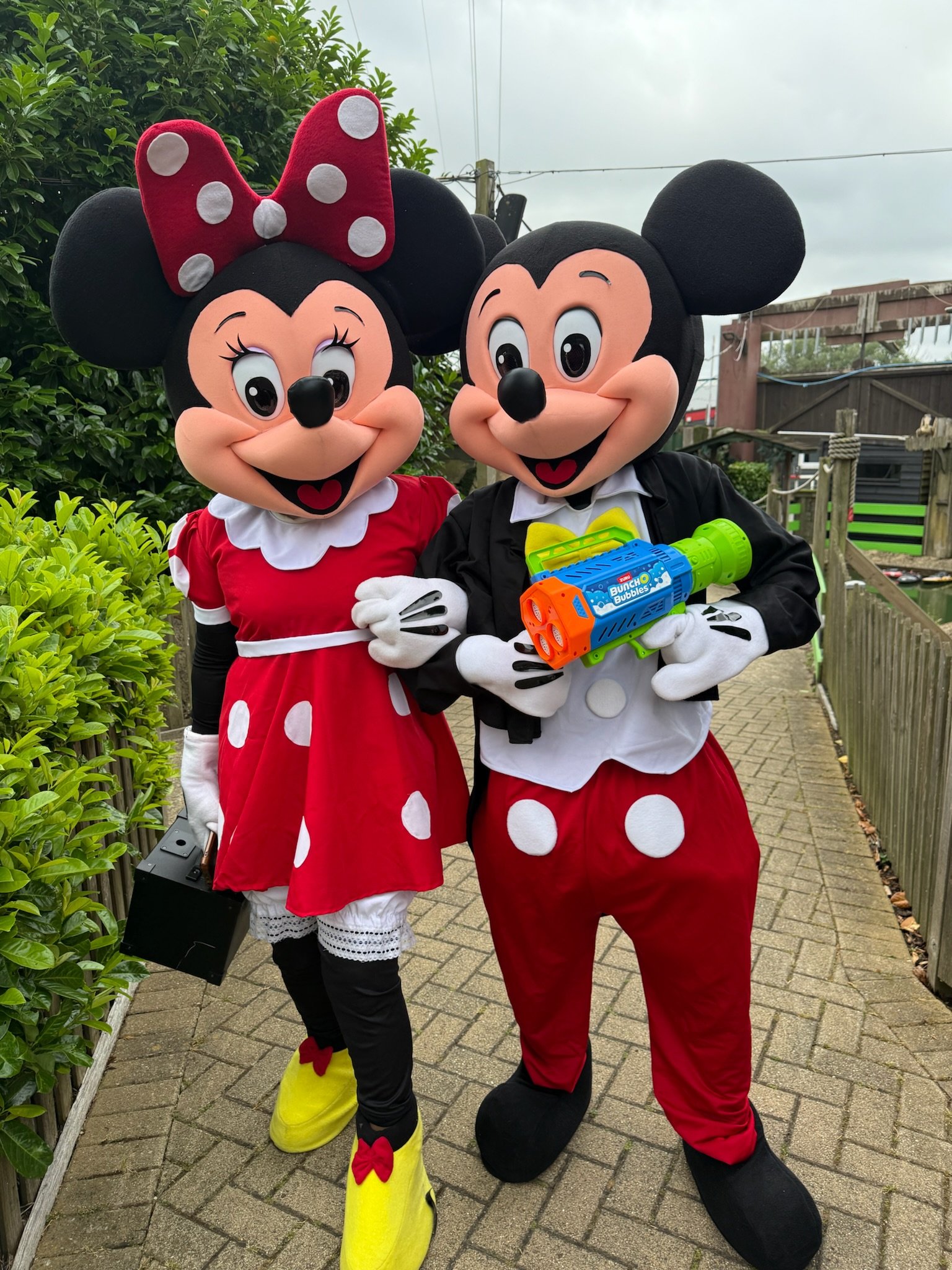 People dressed as Minnie Mouse and Mickey Mouse characters standing outside on a paved path, with greenery and wooden fencing in the background, holding colorful bubble guns.
