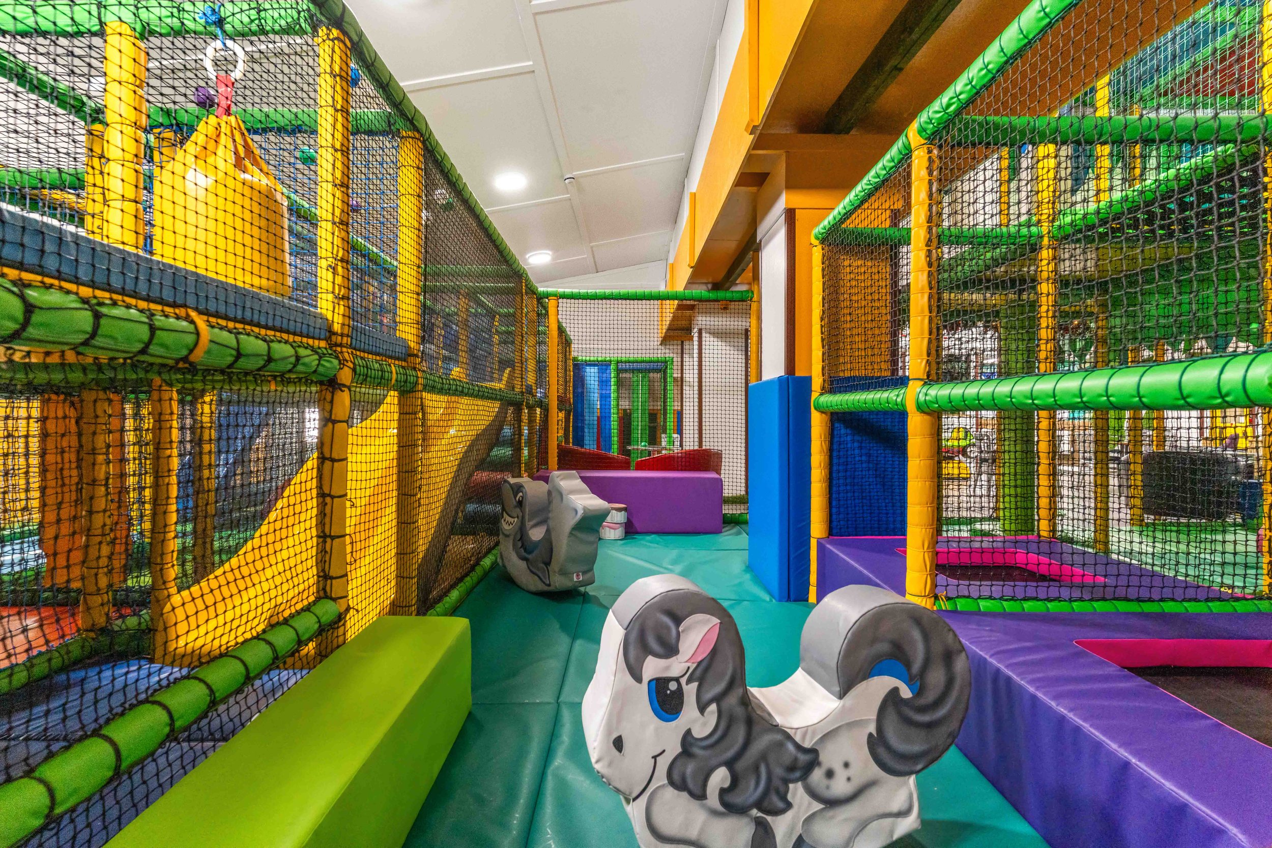 Indoor children's play area with colorful padded structures, slides, and foam animal figures, enclosed by netting.