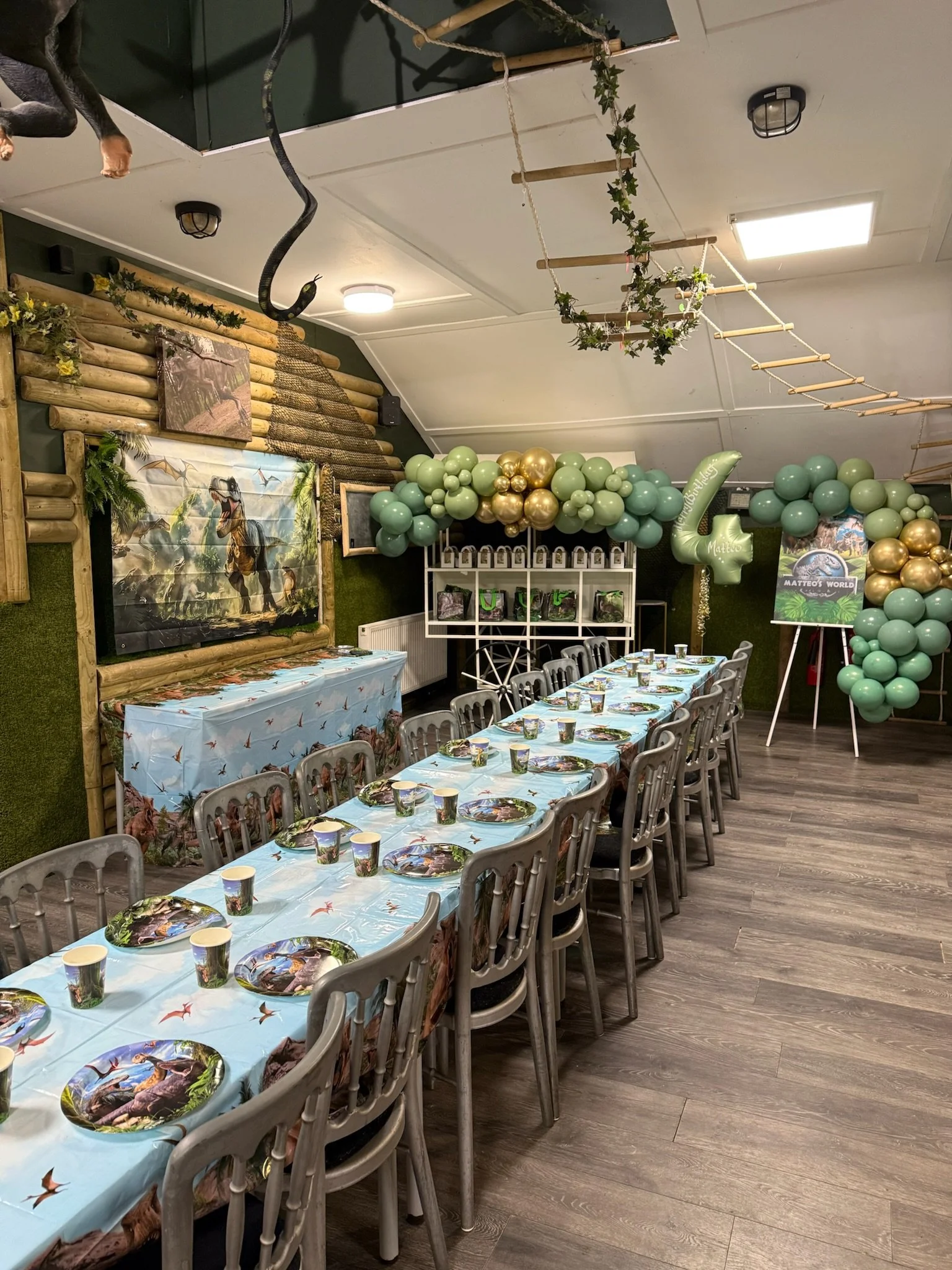 Party room decorated with a dinosaur theme featuring a long table with dinosaur-themed plates and cups, large balloon arrangements in green and gold, a jungle-themed backdrop, and a play ladder, with some wall decorations and a mural of a dinosaur.