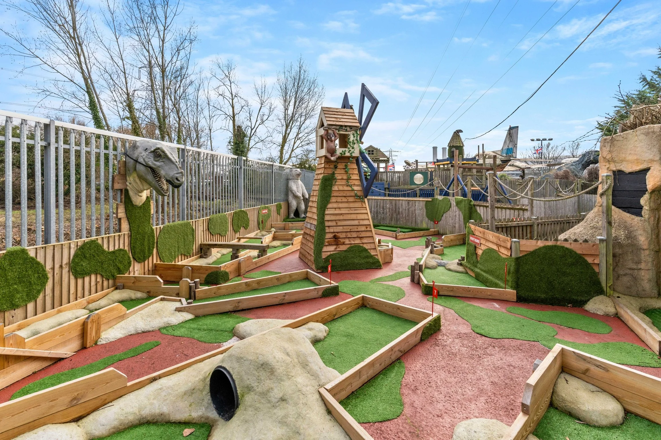Miniature golf course with dinosaur-themed decor, including large dinosaur head on the side, rock formations, artificial grass, and wooden obstacles, set outdoors with trees and cloudy sky in the background.