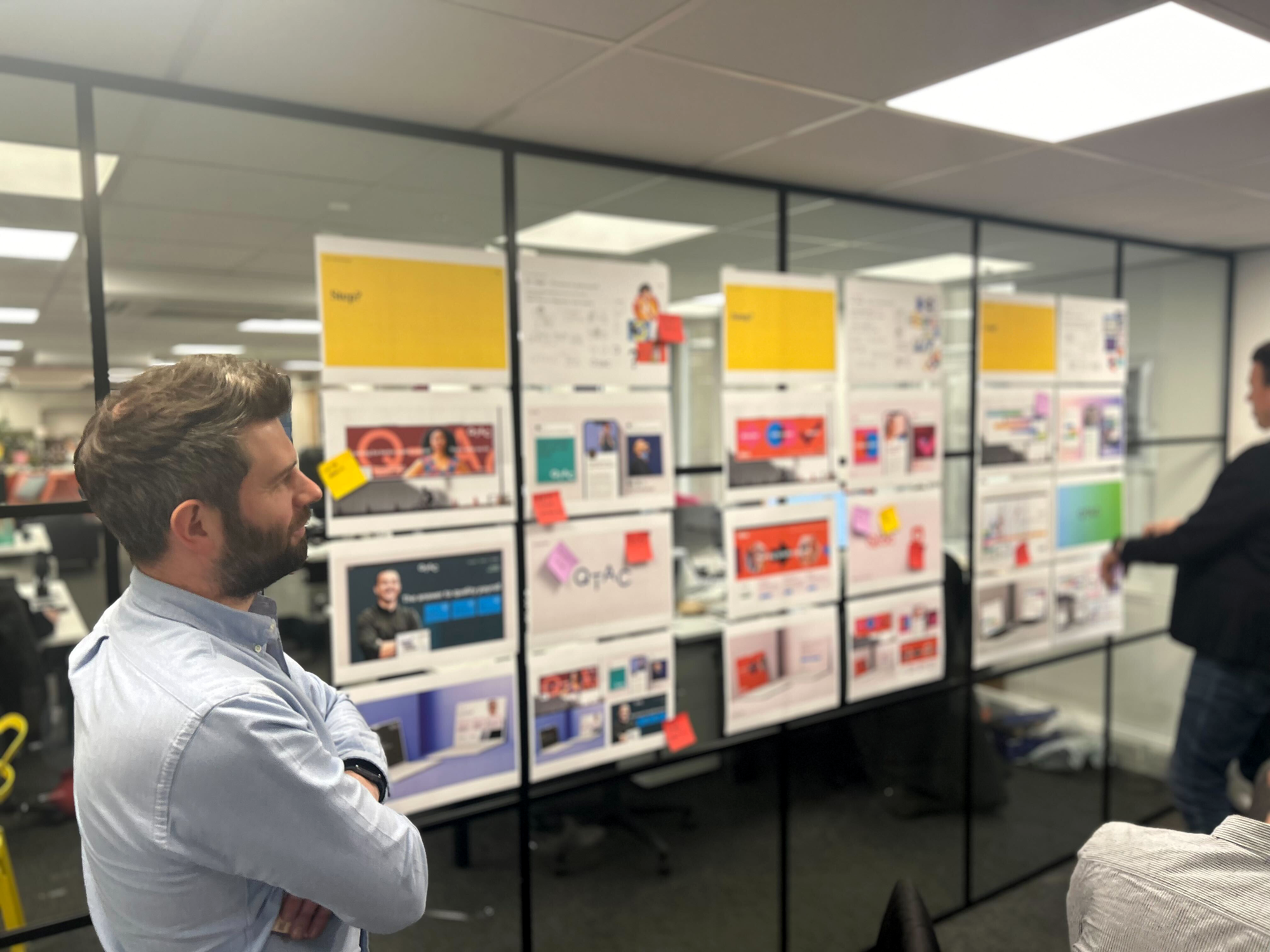 A man with brown hair and a beard in a light blue shirt standing with arms crossed in an office or showroom with display boards of various products and advertisements behind him. It's a brand workshop for discussions around strategy and business