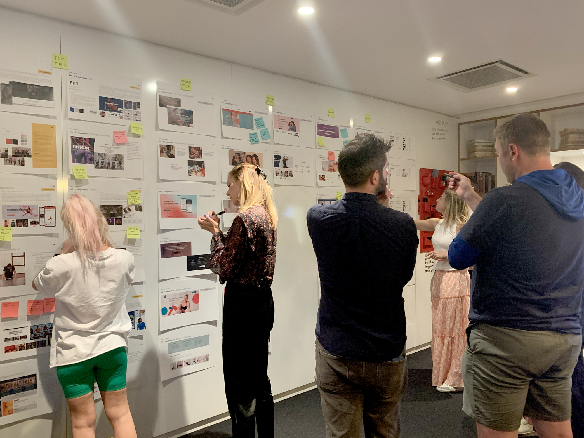 People gather around a wall with various sheets of paper, sketches, and sticky notes in an office or collaborative workspace. Some individuals are examining the documents, and one is using a tool or pen to point at the wall.