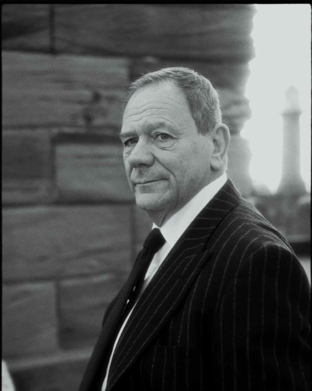 A black and white portrait of a middle-aged man with short hair, wearing a pinstripe suit and tie, standing outdoors with a wooden structure and a chimney in the background.