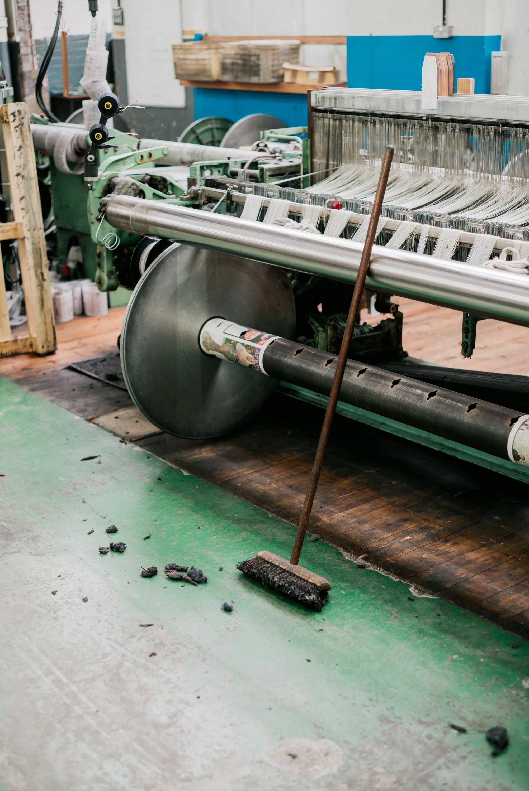 A cleaning broom leaning against a large industrial machine in a workshop or manufacturing setting. There is debris on the floor and a mop underneath the machine.