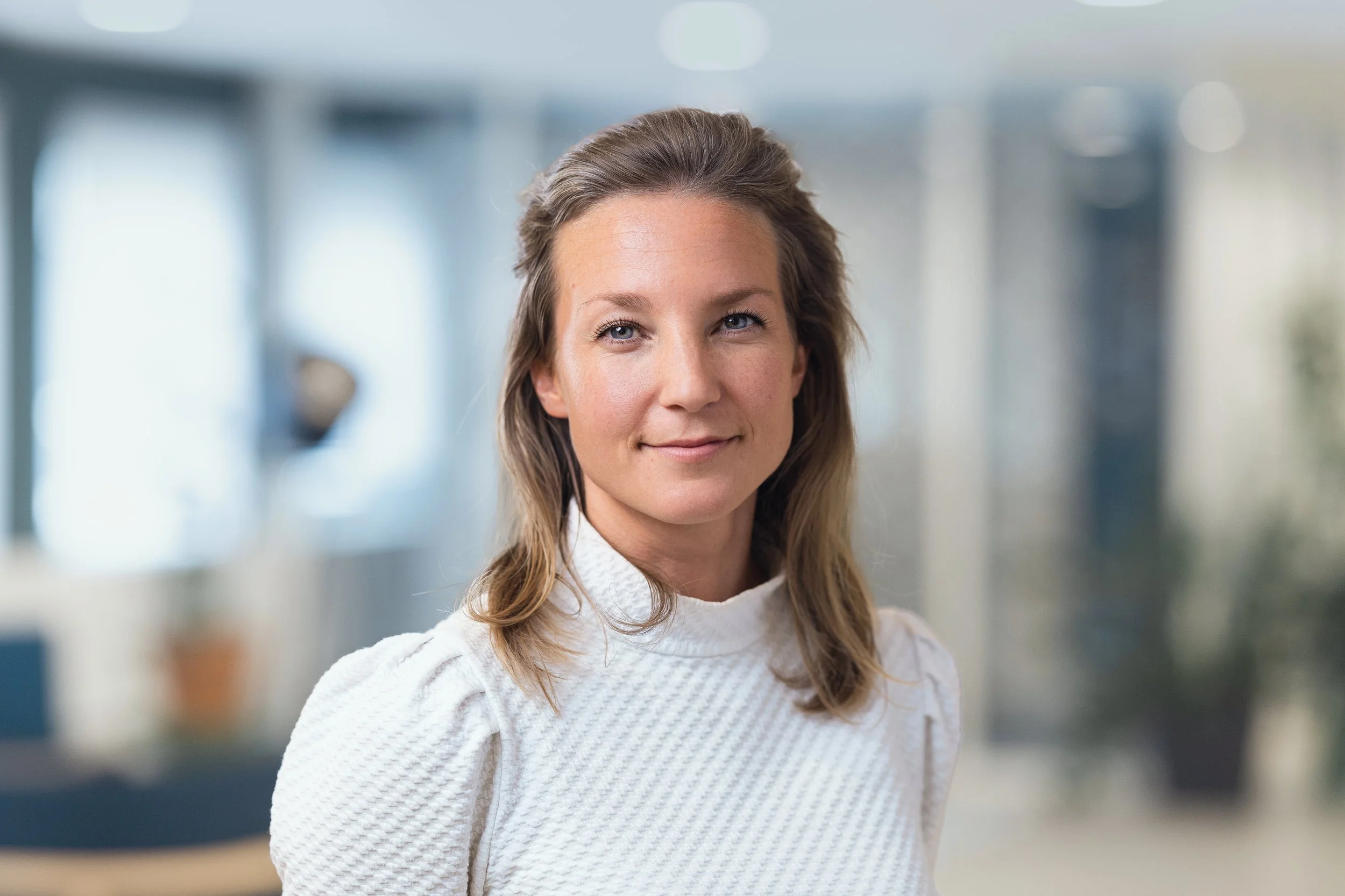 Portret van een jonge vrouw met halflang, golvend bruin haar in een lichte kantooromgeving.