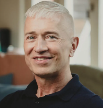 Portret van een man met kort, wit haar die glimlacht in een interieur met zachte verlichting.