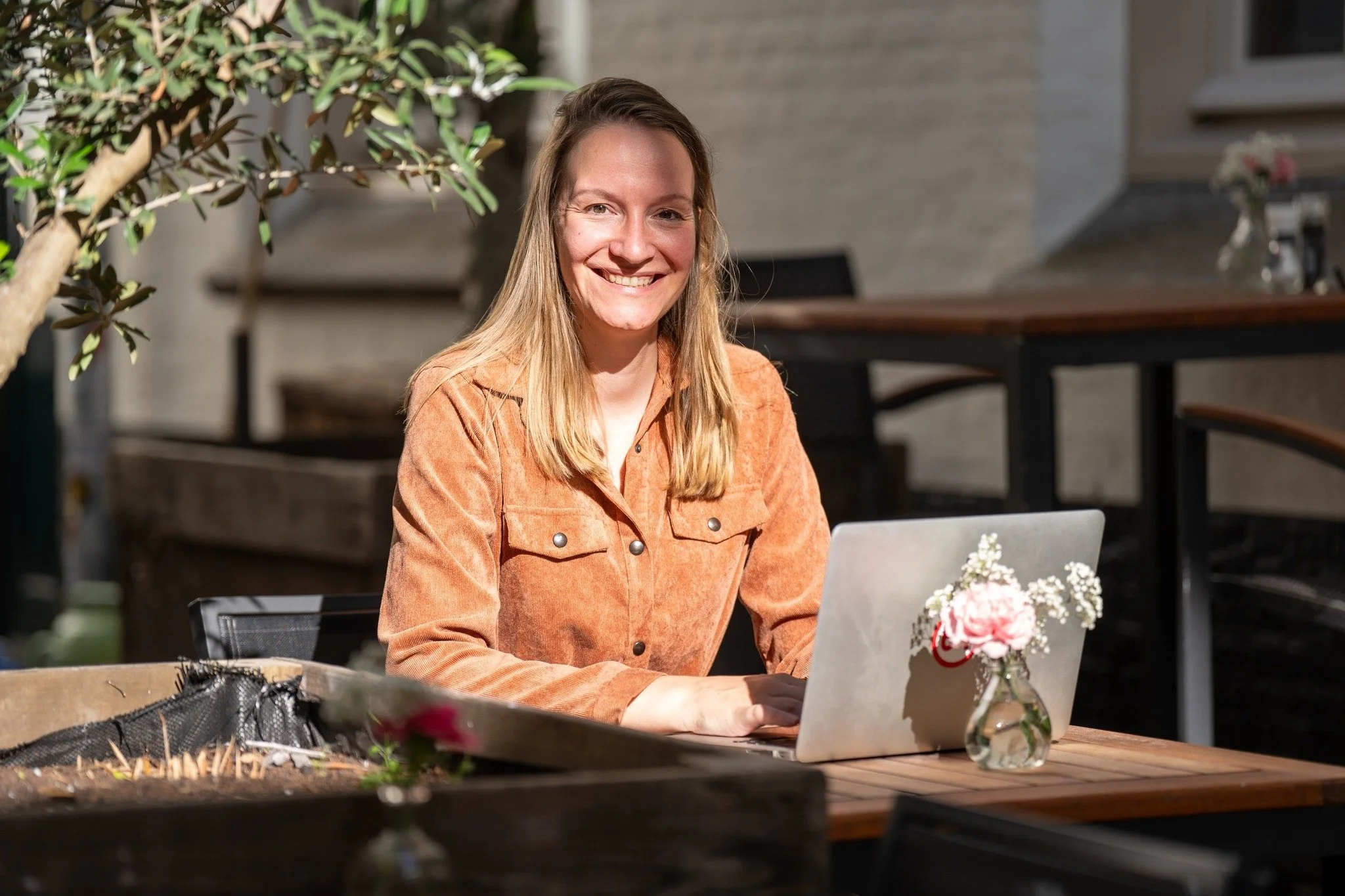 Vrouw met lange blonde haar zit buiten aan een tafel met haar laptop, lachend naar de camera. Op de tafel staat een klein vaasje met bloemen.