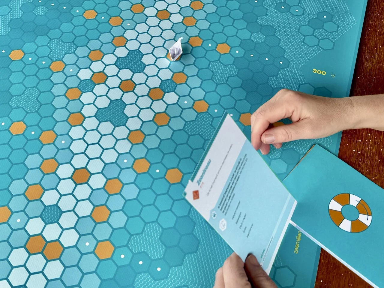 Een bordsspel Het Boordspel met hexagonale tegels in blauw en oranje. Een hand houdt een kaart vast, en er ligt een kaart met een reddingsboei erop.