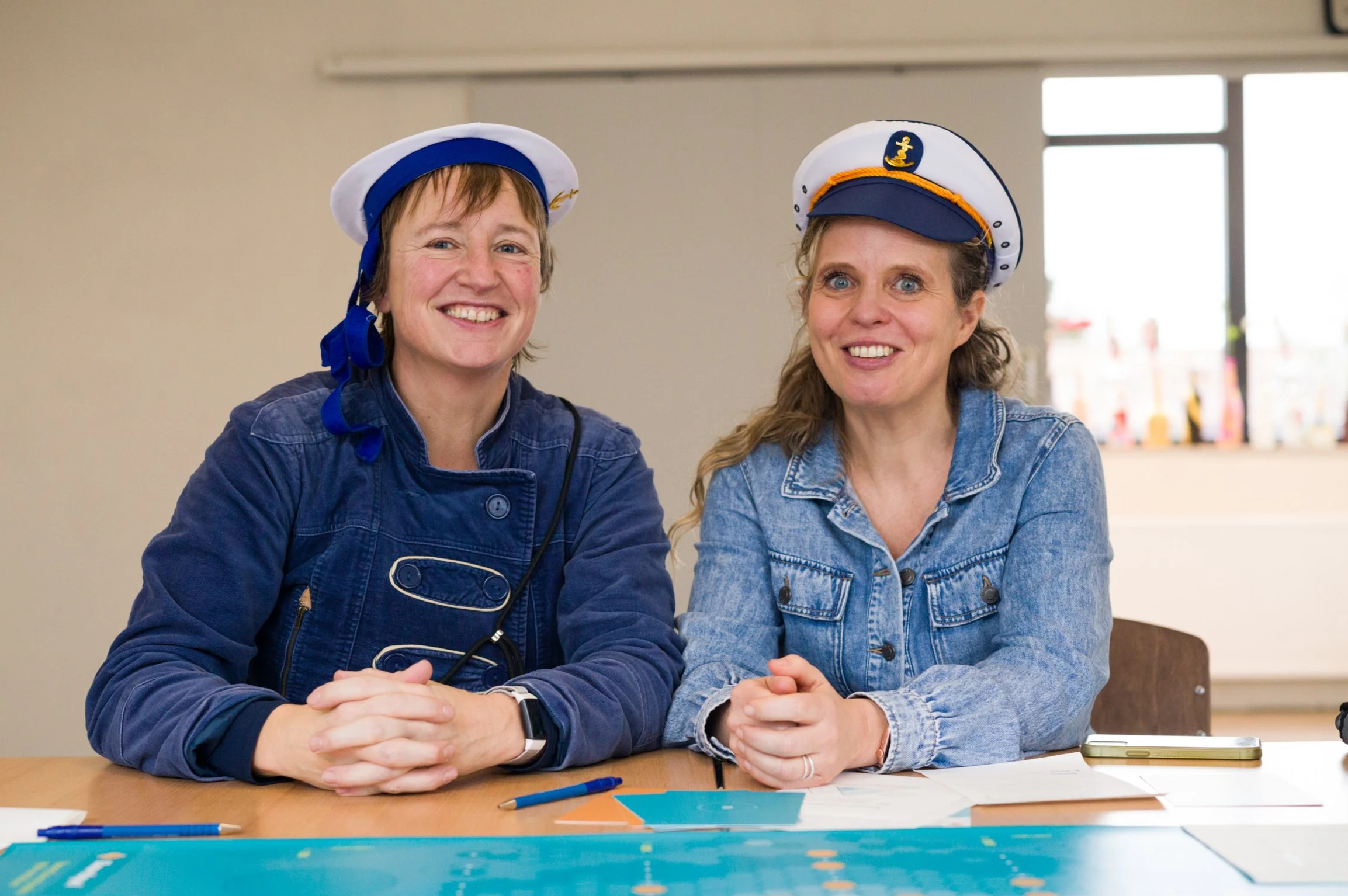 Twee vrouwen, de bedenkers van het Boordspel, hebben een kapiteinspet op en zitten aan een tafel met papieren en pennen, lachend en voor de camera. Voor hun het Boordspel.