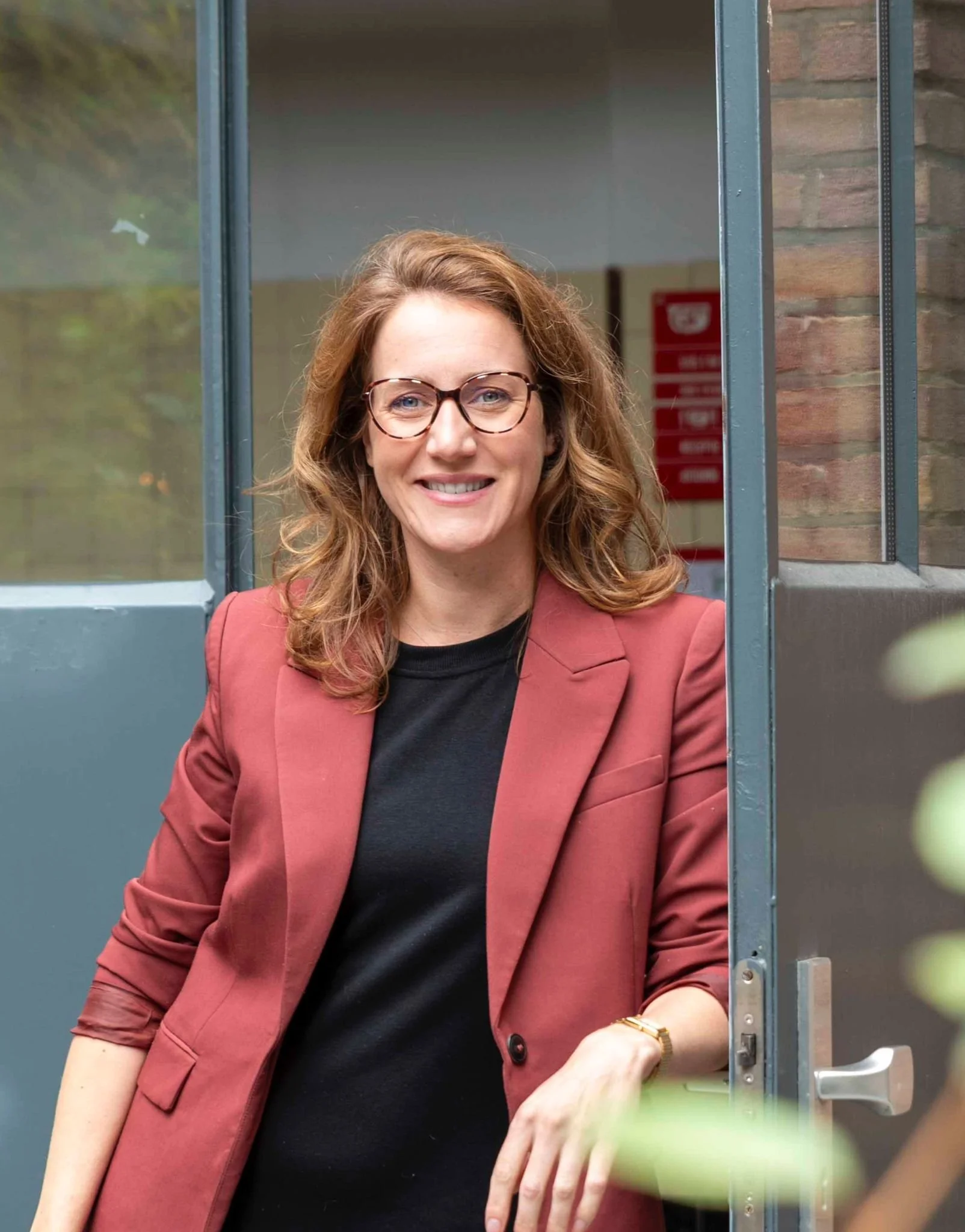 Een vrouw met bruin haar en bril staat bij een deur, lachend naar de camera.