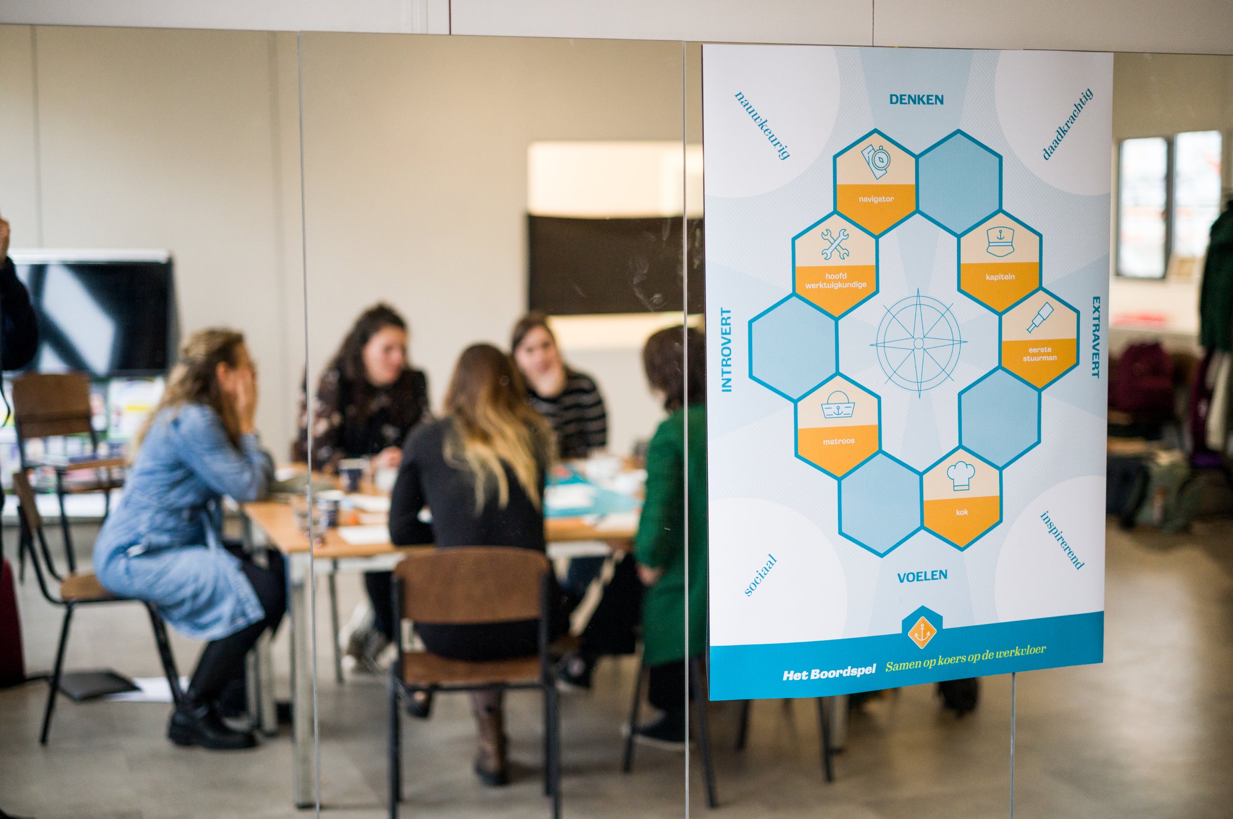 Mensen die in een vergaderruimte bij een tafel zitten, met een poster over teamrollen en communicatie op de voorgrond.