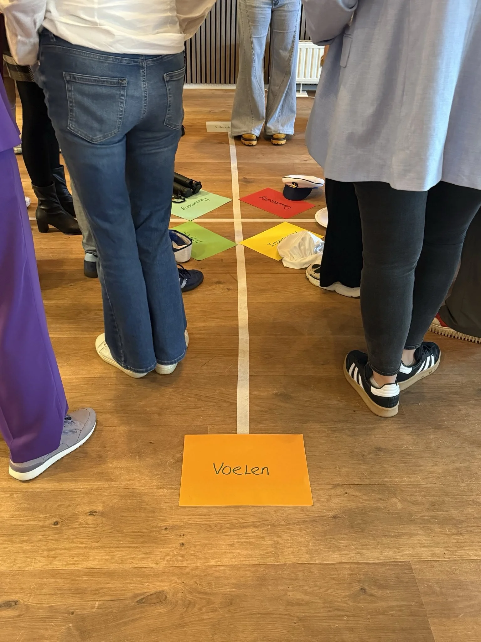 De afbeelding toont mensen die staan rond een vloer met gekleurde kaarten en papieren, met een oranje kaart waarop 'Voelen' staat. Onderdeel van teamwiel DISC.