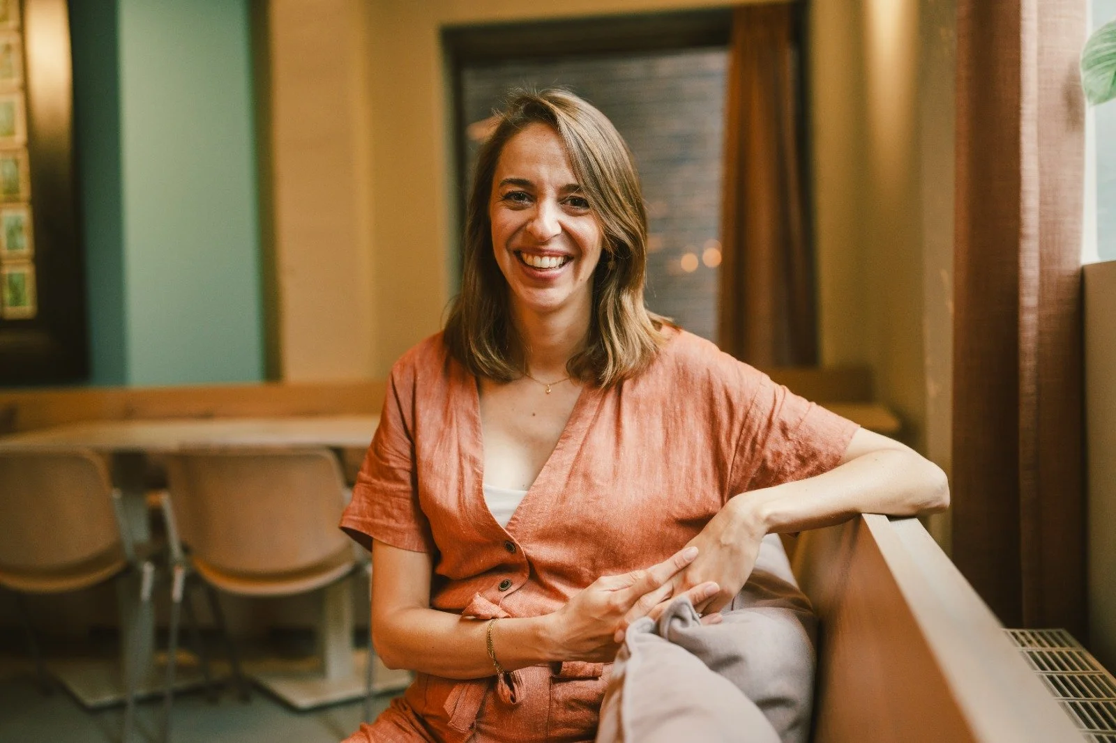 Een lachende vrouw met schouderlang bruin haar zit op een bank in een gezellige ruimte, gekleed in een oranje blouse.