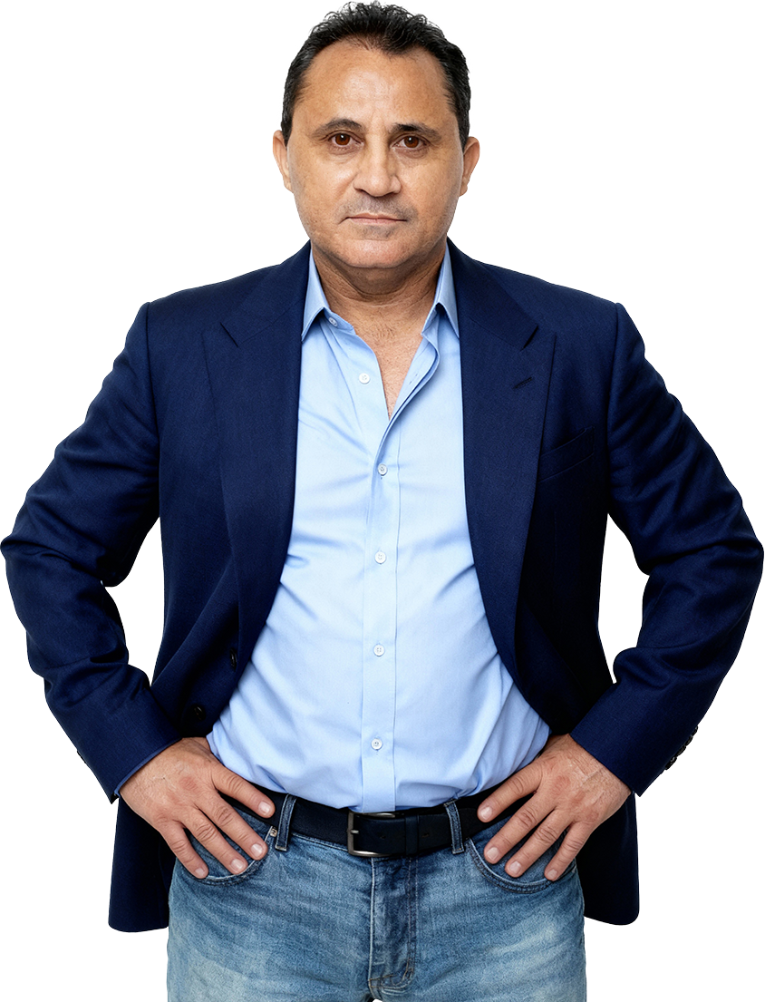 Homem de meia idade, com cabelo escuro e pele clara, usando um blazer azul escuro, camisa azul clara e jeans, com as mãos apoiadas na cintura, olhando seriamente para a câmera.