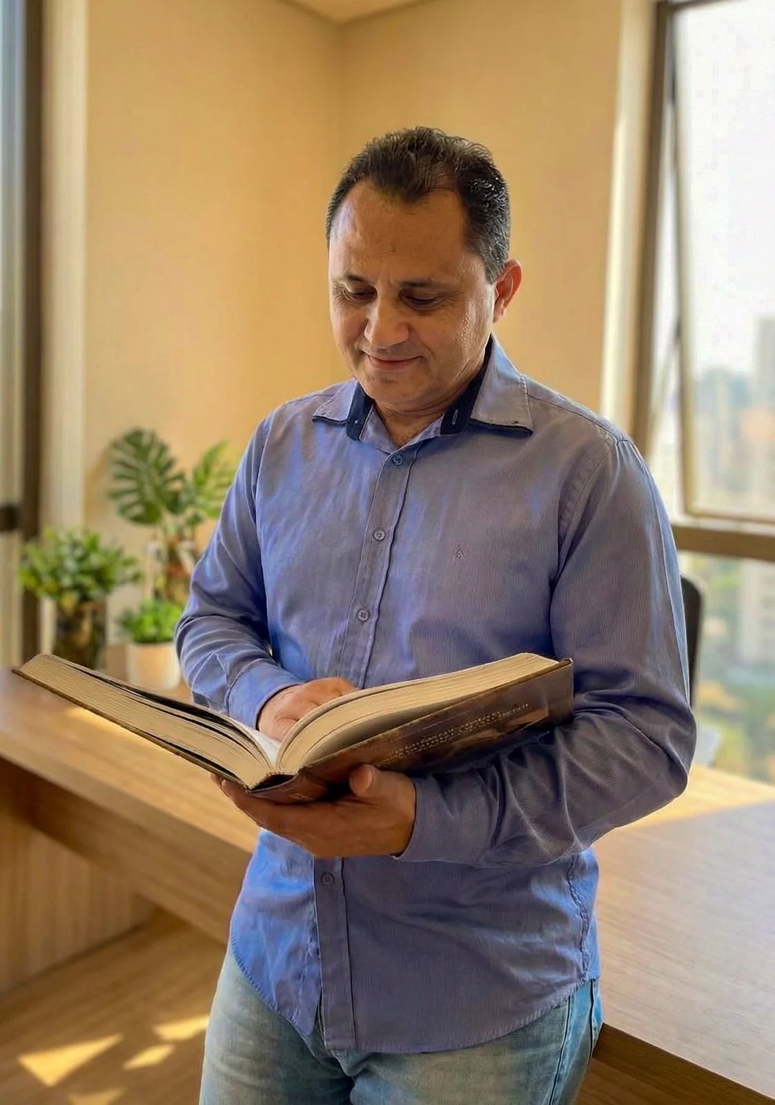 Homem lendo um livro em um ambiente bem iluminado com plantas ao fundo.