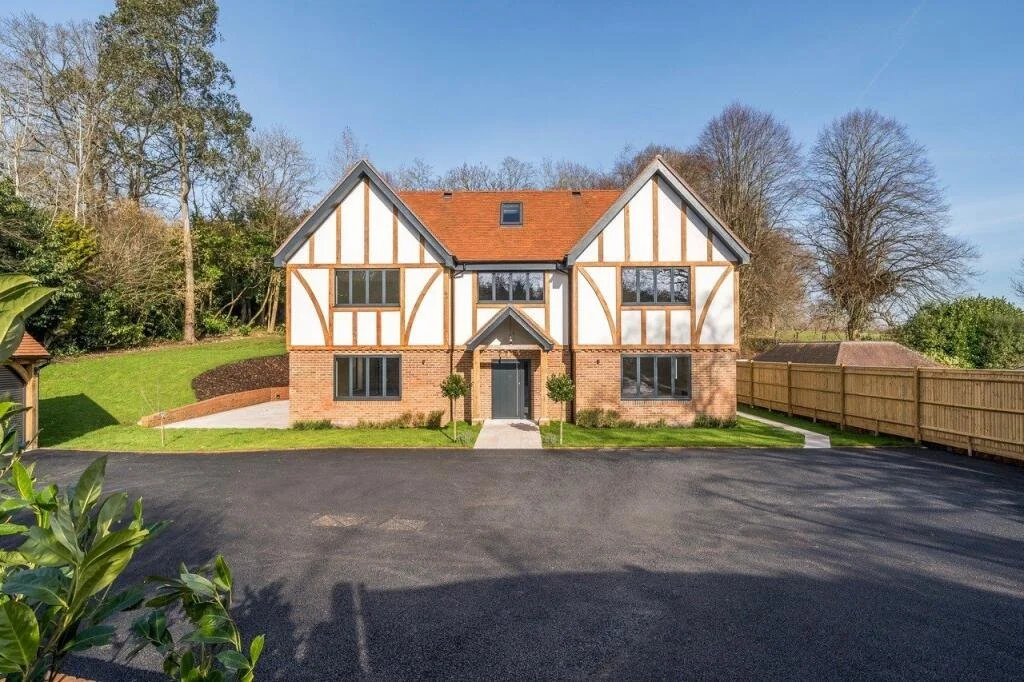 A two-story house with half-timbered design and brick foundation, surrounded by a green lawn, trees, and a paved driveway.