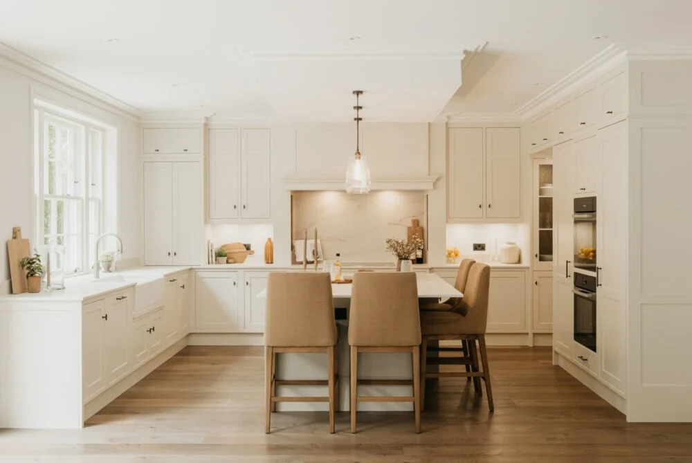 Bright kitchen with white cabinetry, beige chairs around a central island, and a window with natural light.
