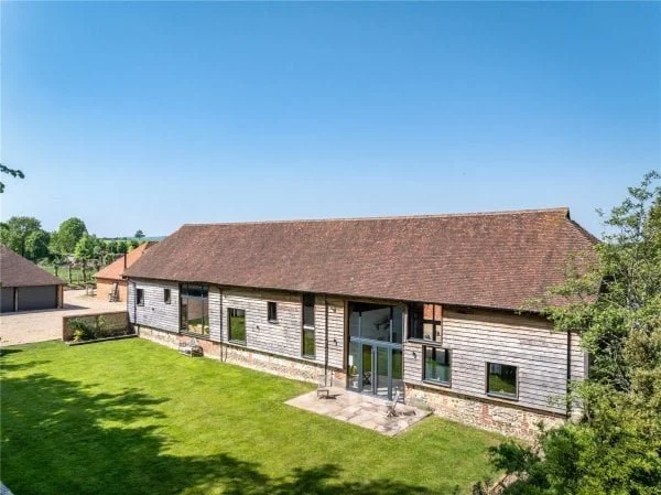 A large, single-story house with a brown tiled roof, wooden siding, and multiple windows, surrounded by a green lawn and trees, under a clear blue sky.