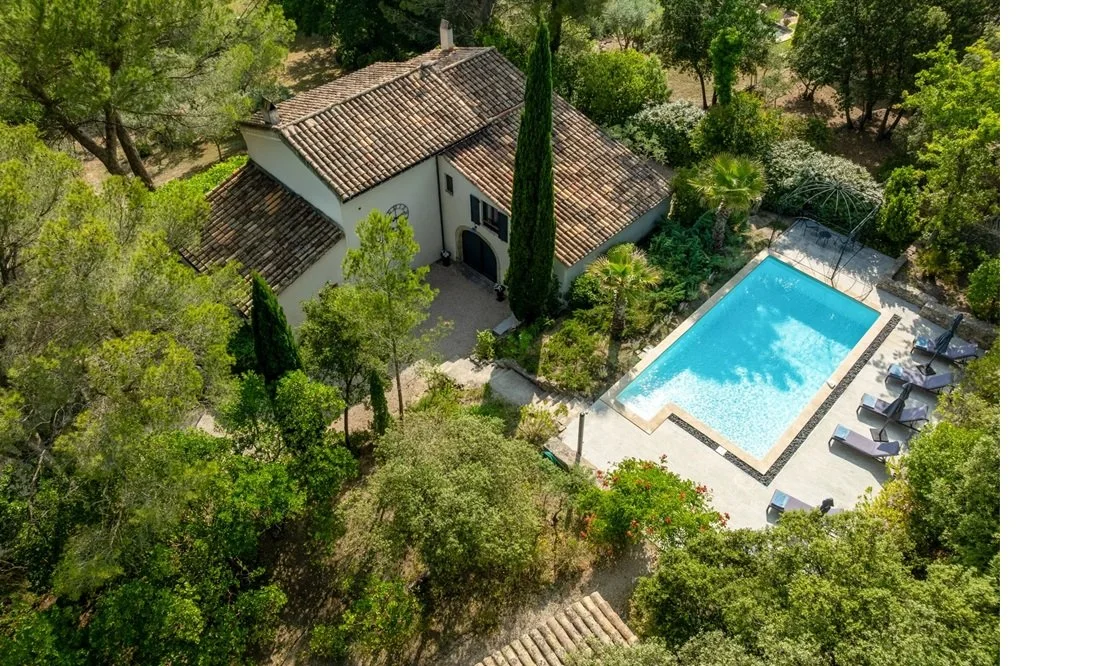 Aerial view of a house surrounded by trees with a swimming pool and several lounge chairs nearby.