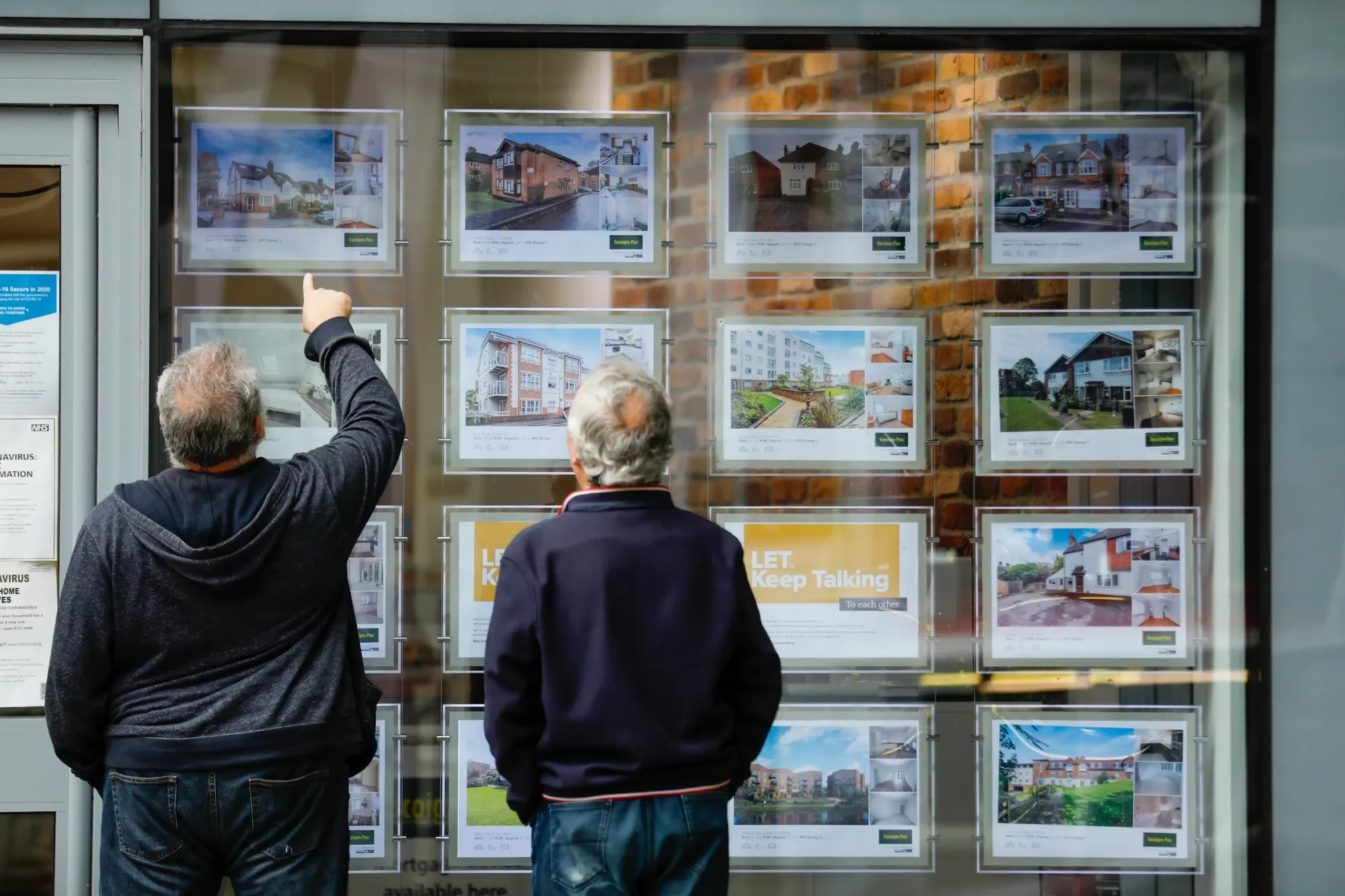 Two people standing outside a real estate window display looking at house listings, with one person pointing at the listings.
