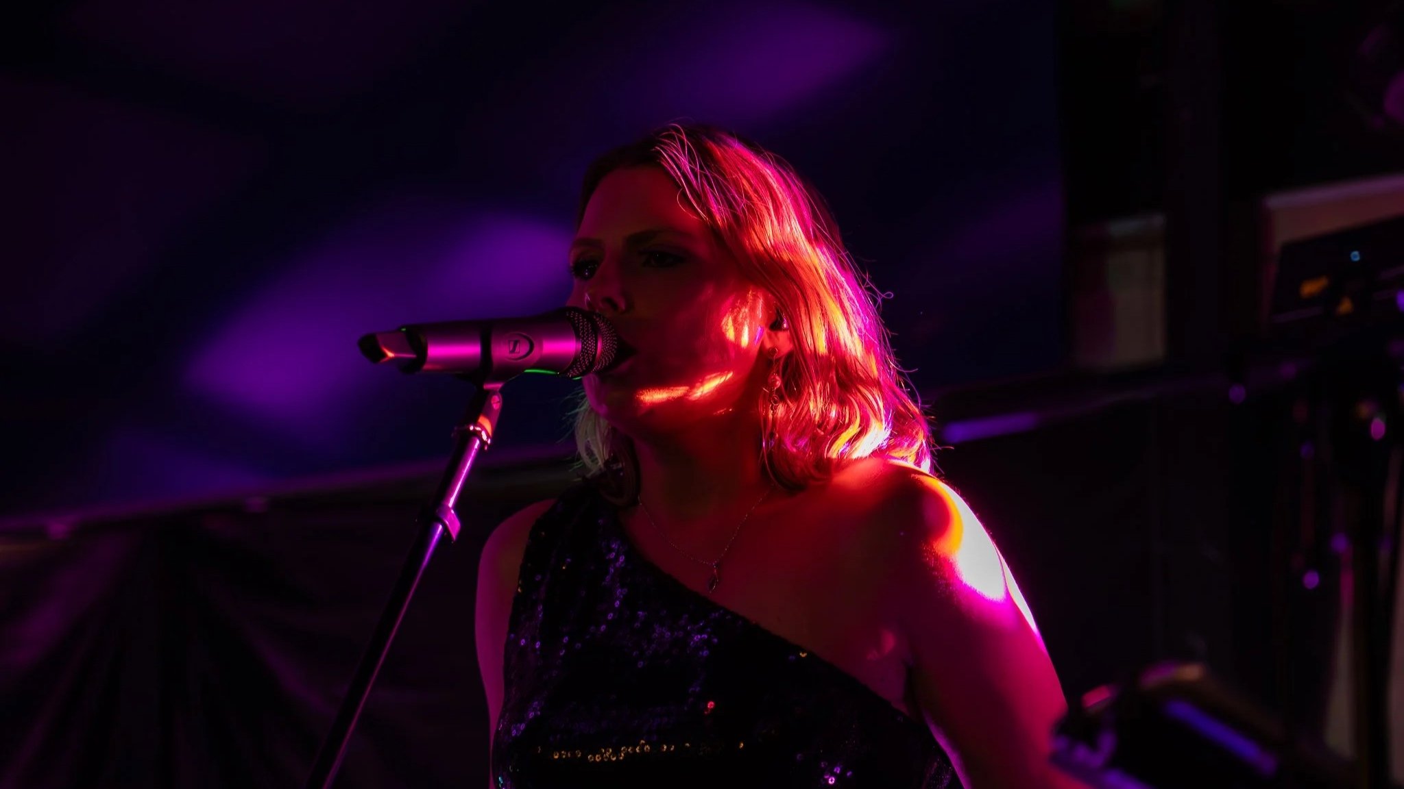 A woman singing into a microphone on stage, illuminated by pink and purple stage lights, with a dark background.