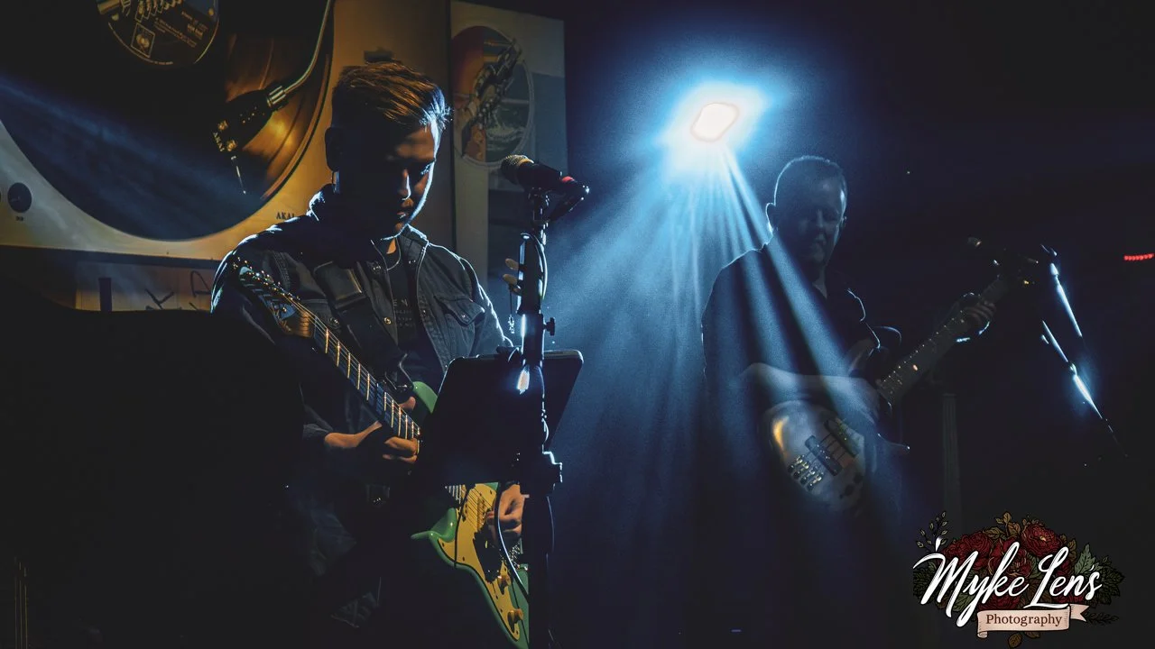 Two musicians playing guitars on a dark stage, illuminated by blue and white stage lights, with a microphone in front of them.