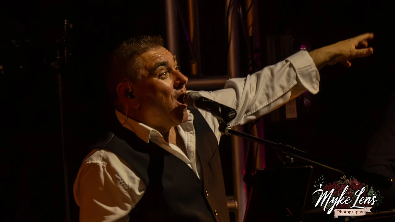 A male singer performing on stage, singing into a microphone, with his arm outstretched. Dark background with stage lights and equipment.