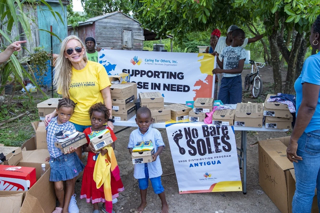 Volunteers distribute shoes to children in a rural area for a charitable event supporting a shoe distribution program in Antigua. Children hold new shoes, and boxes of shoes are on tables.