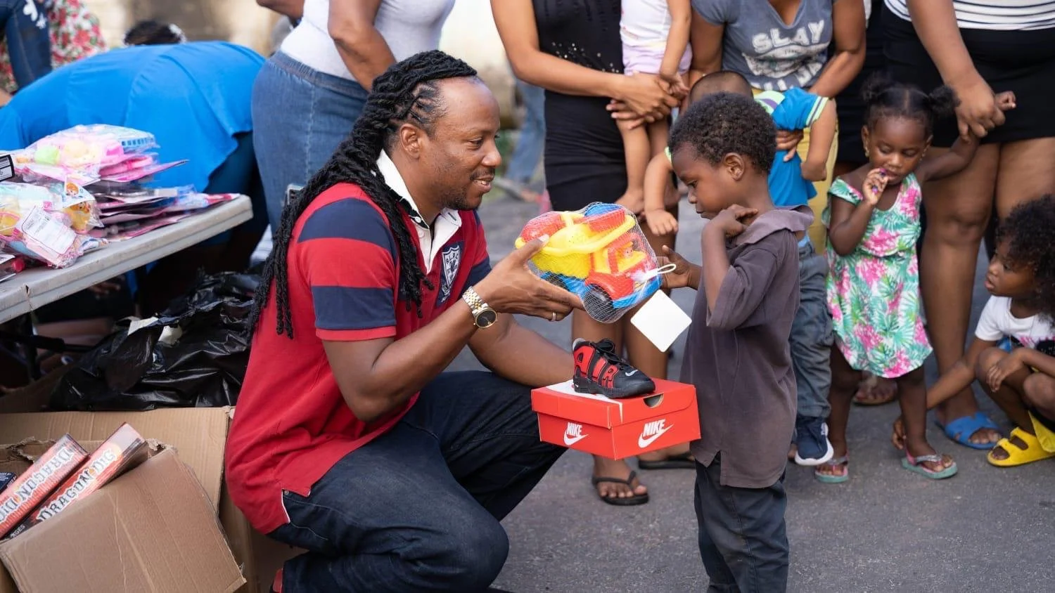 A man with dreadlocks in a red and navy striped polo shirt is kneeling and handing a boxed Nike shoe to a young boy. The boy is receiving a toy set in a mesh bag from the man. Several children and adults are gathered around, some standing, some sitting, with a table of toys and supplies behind them.