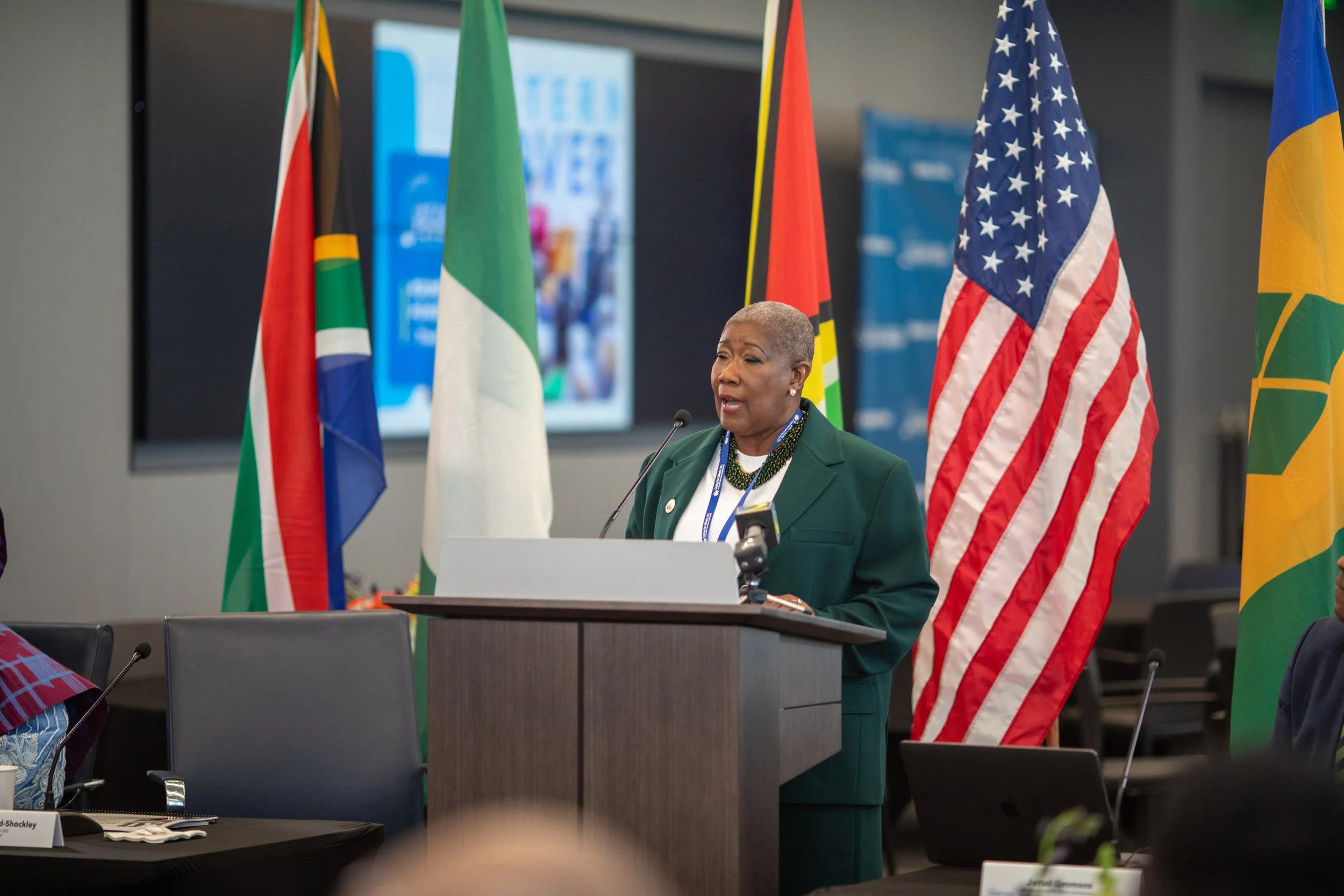 A woman with short gray hair giving a speech at a podium with flags of different countries, including the United States, South Africa, Italy, and Brazil, in the background.