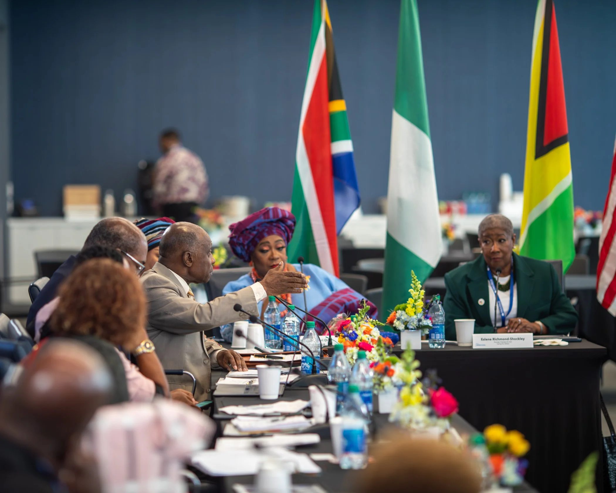 A diverse group of people sitting at a conference table with flags in the background, engaging in discussion. Items like water bottles, papers, and flowers are on the table.