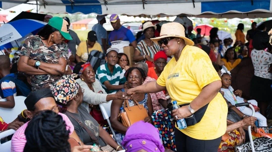 A woman in a yellow shirt and straw hat shaking hands with an older woman seated under a tent at a crowded outdoor event, with multiple people sitting and standing around.