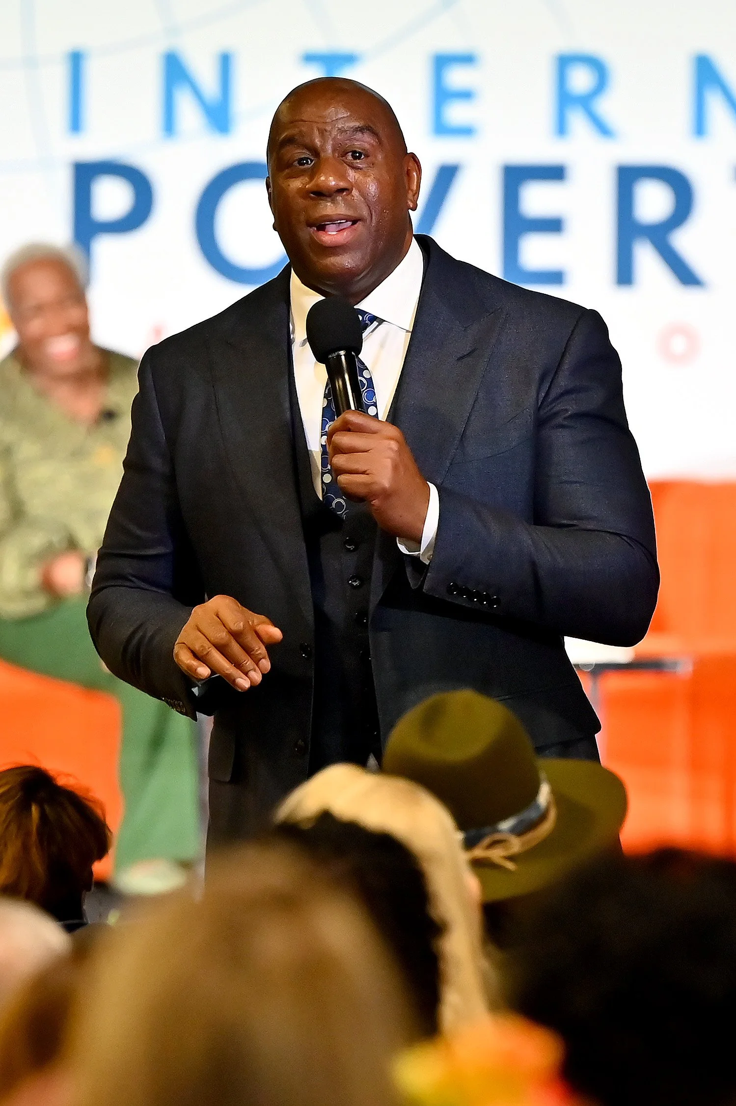 A man in a dark suit speaking into a microphone at an event, with a woman smiling in the background and a partially visible audience.