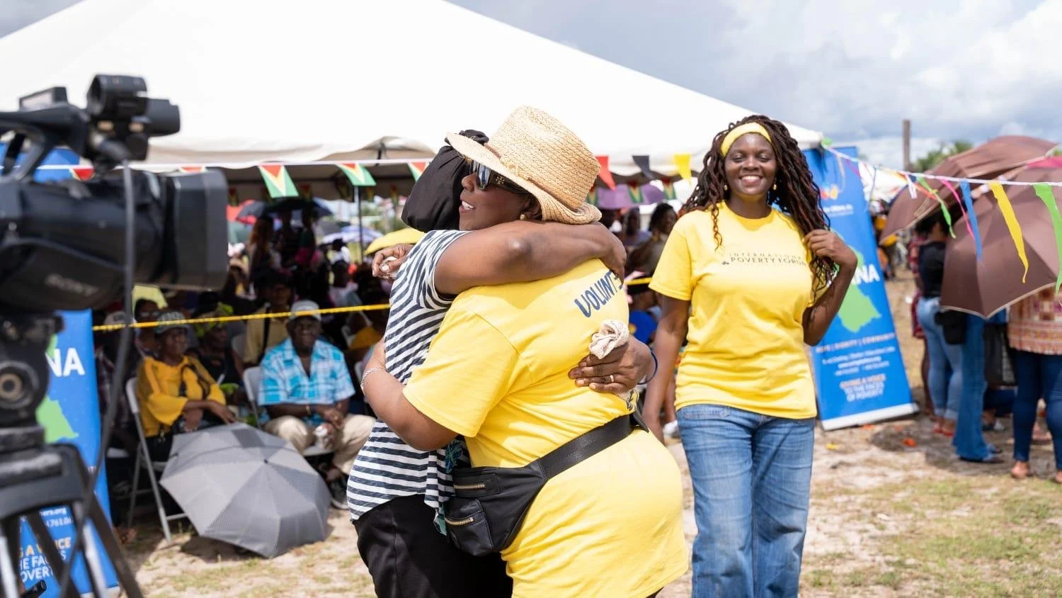 Two women hugging at an outdoor event, surrounded by people, tents, and banners.