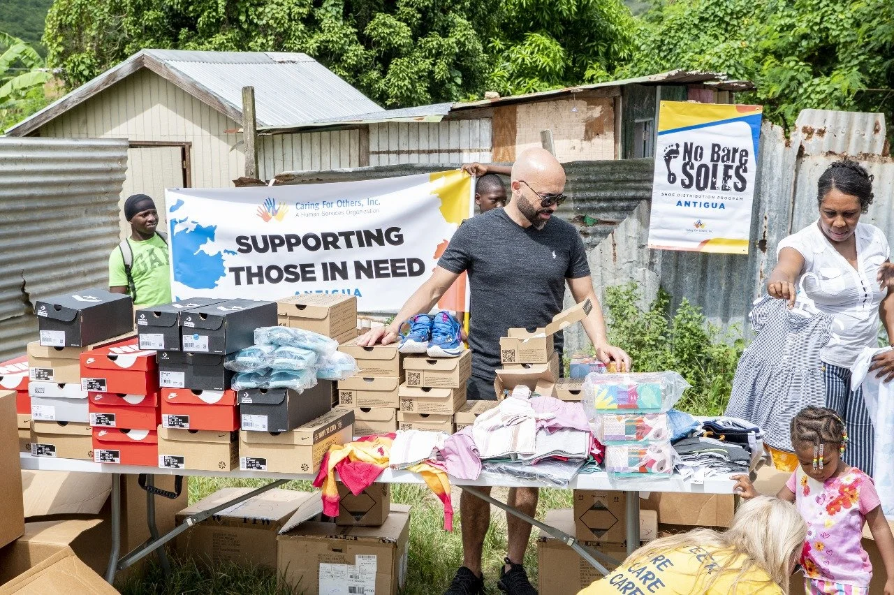 People participating in a community shoe distribution event outdoors, with tables of shoes and clothing, and banners reading supporting those in need and No Bare Soles.