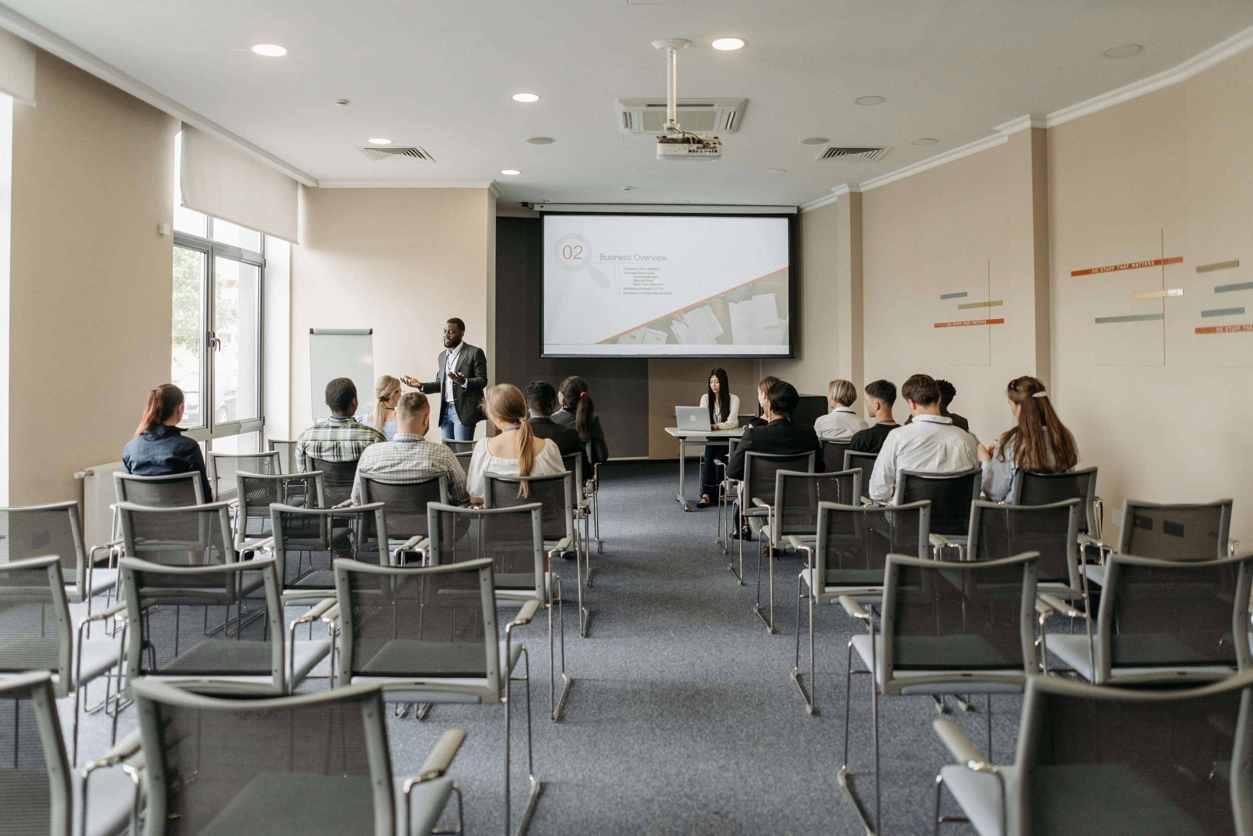 Business meeting or presentation in a conference room with a speaker at the front and an audience seated facing him, on a large projector screen.