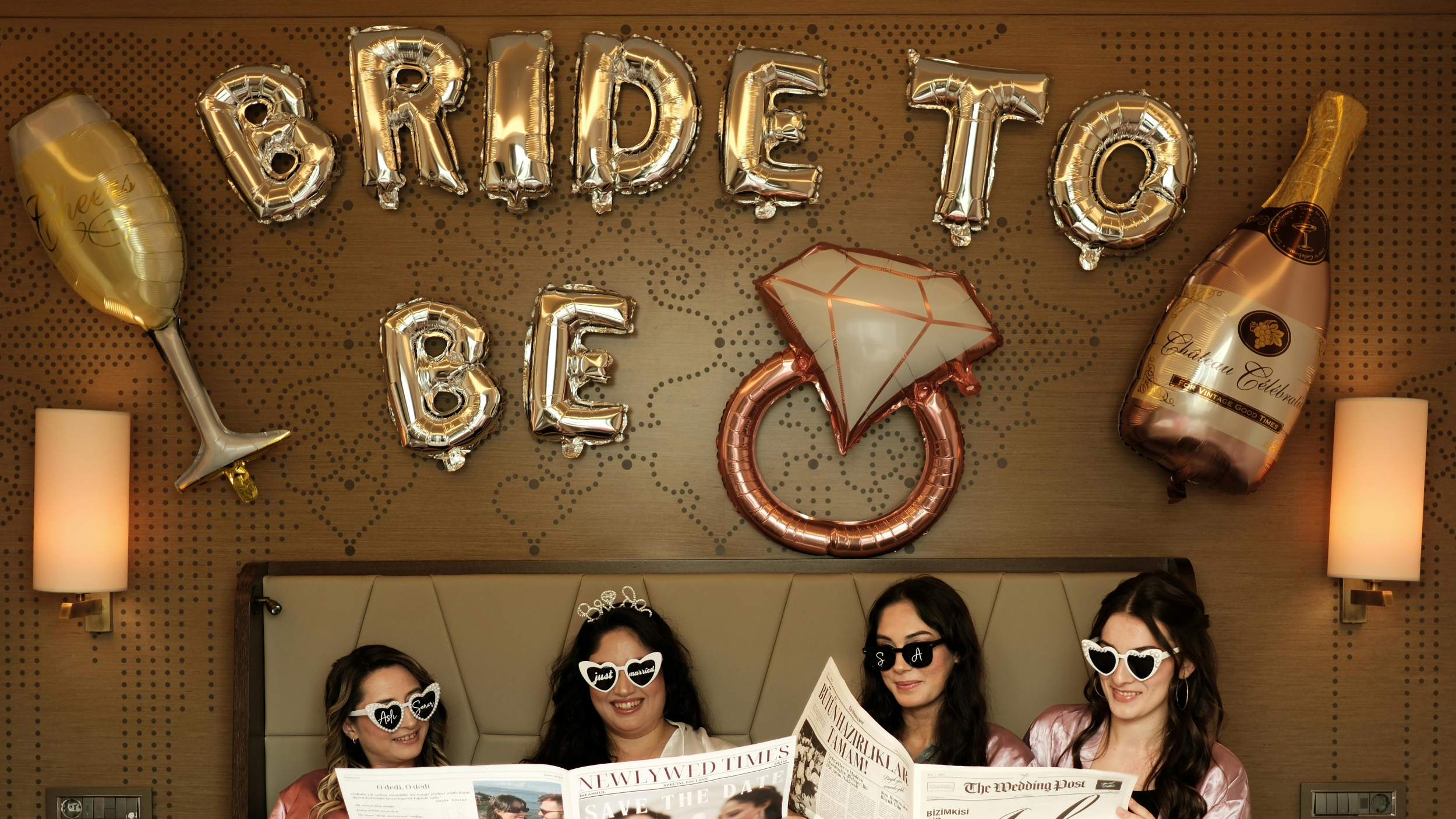 Four women sitting in bed, reading newspapers, celebrating a bridal shower with metallic letter balloons that spell 'BRIDE TO BE' and shaped balloons of a diamond ring, champagne bottle, and champagne glass, with wall-mounted lights in a decorated room.